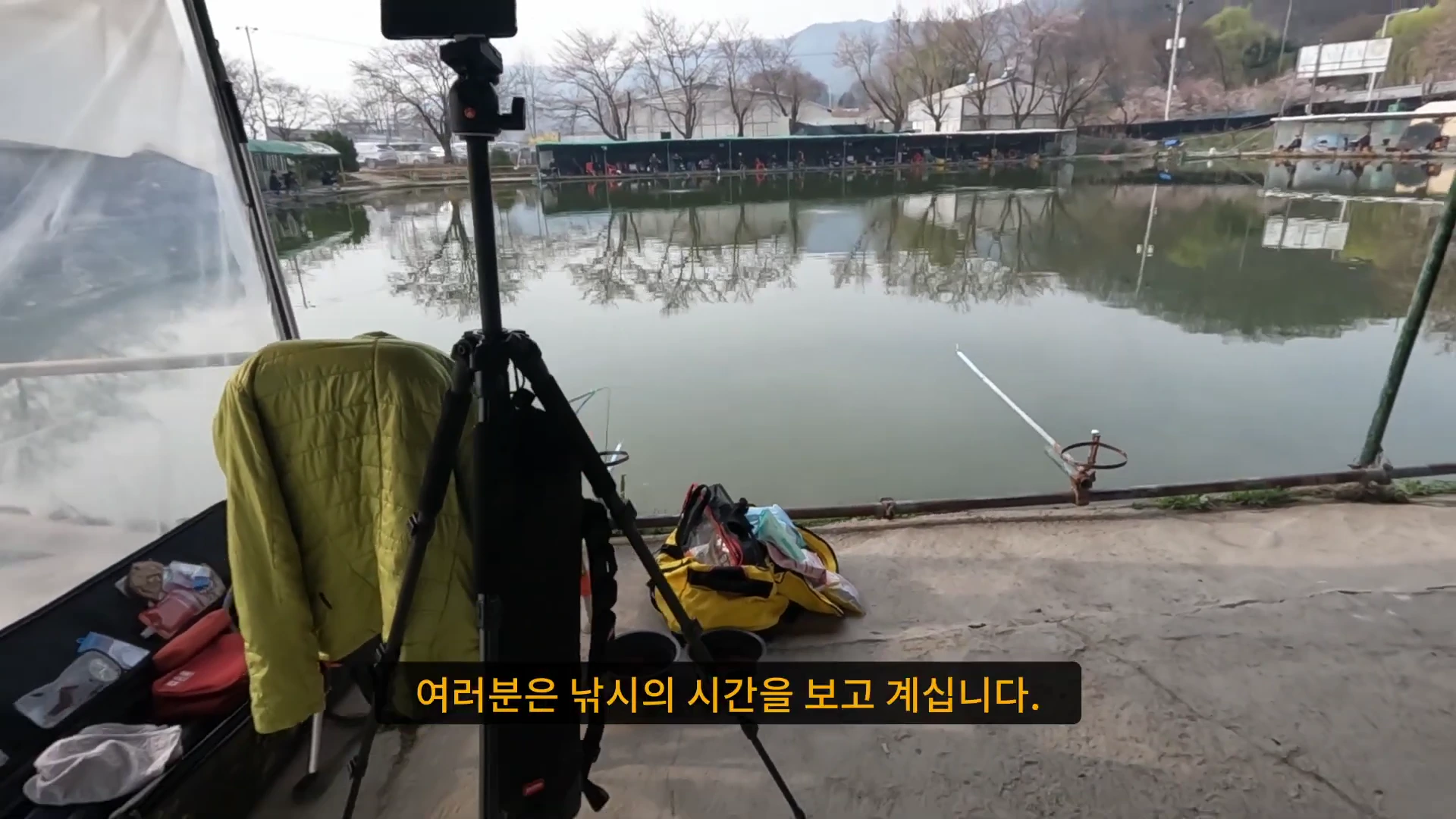 낚시터 의자에 카메라 삼각대가 세워져 있고 앞쪽으로 호수와 맞은편 낚시 좌대가 보이는 풍경.