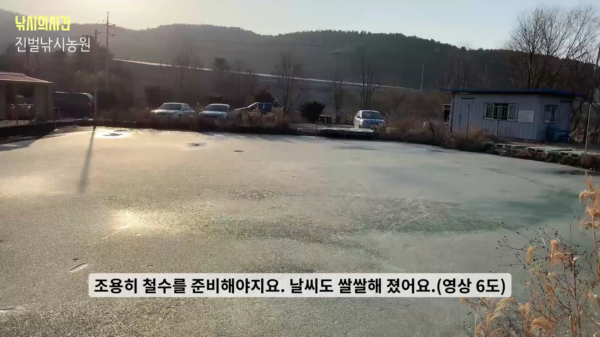 얼어붙은 낚시터 수면 위로 겨울 햇살이 비치고 있으며 뒤편에는 주차된 차량들과 관리 건물이 보이는 풍경