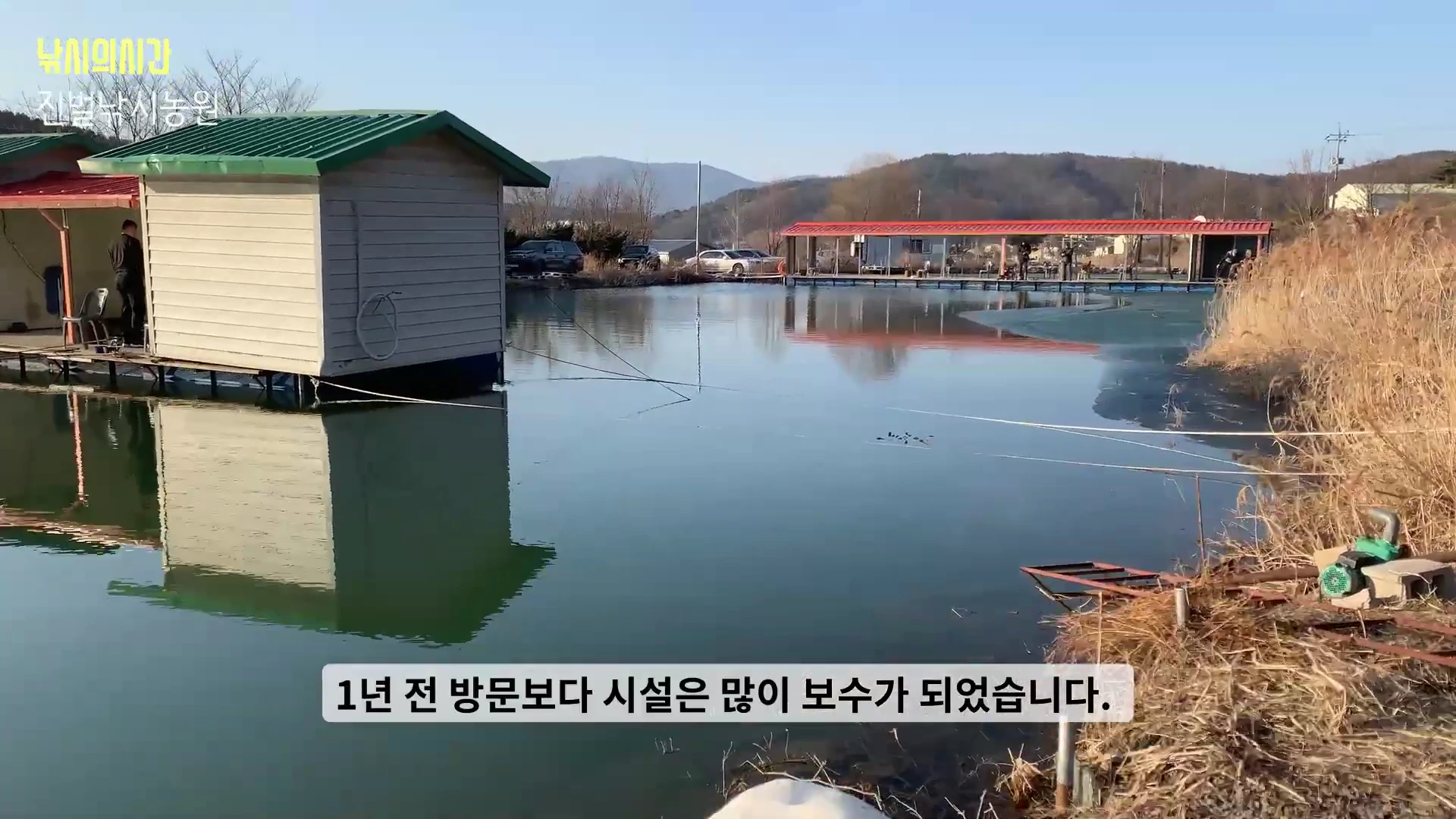 낚시터의 전경을 담은 영상으로, 연못 위에 떠 있는 작은 방갈로와 맞은편으로 길게 이어진 낚시 좌대가 보입니다.