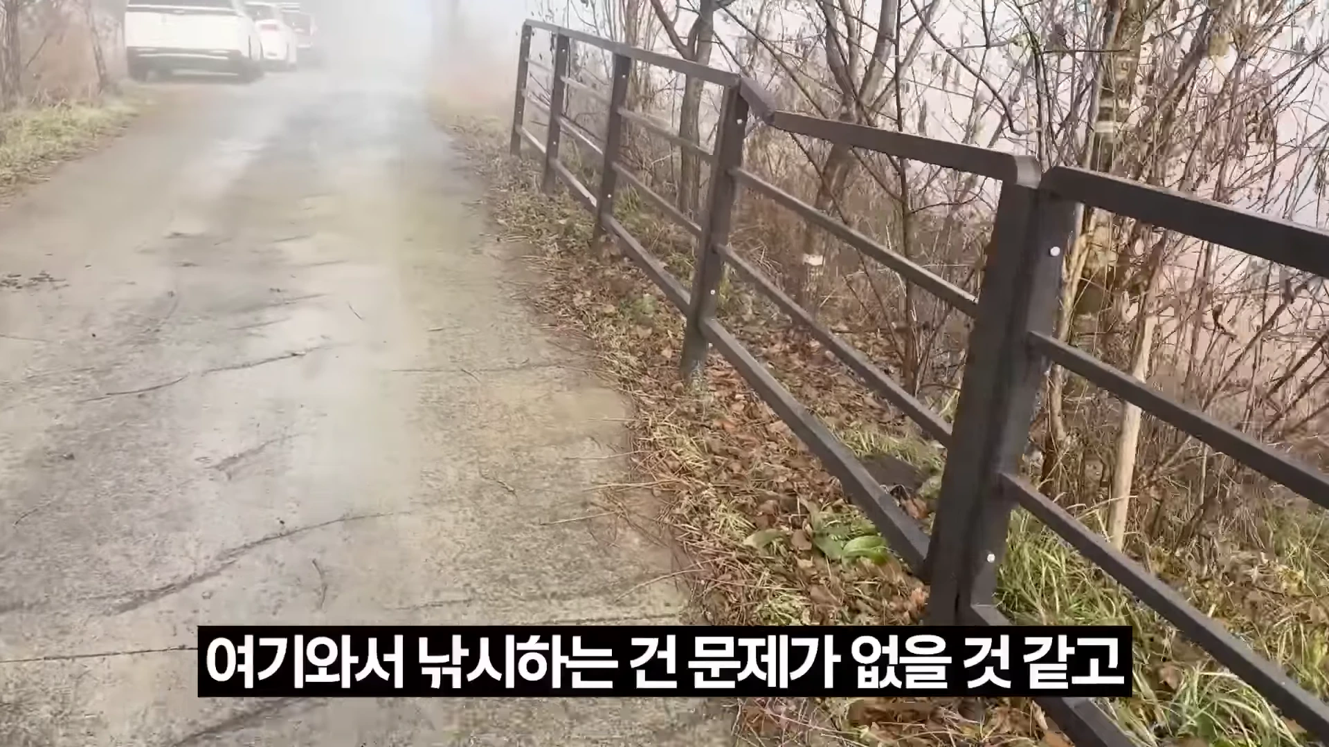 안개 낀 강변의 비포장도로 옆으로 금속 울타리가 길게 이어져 있고, 멀리 차량들이 주차되어 있는 모습