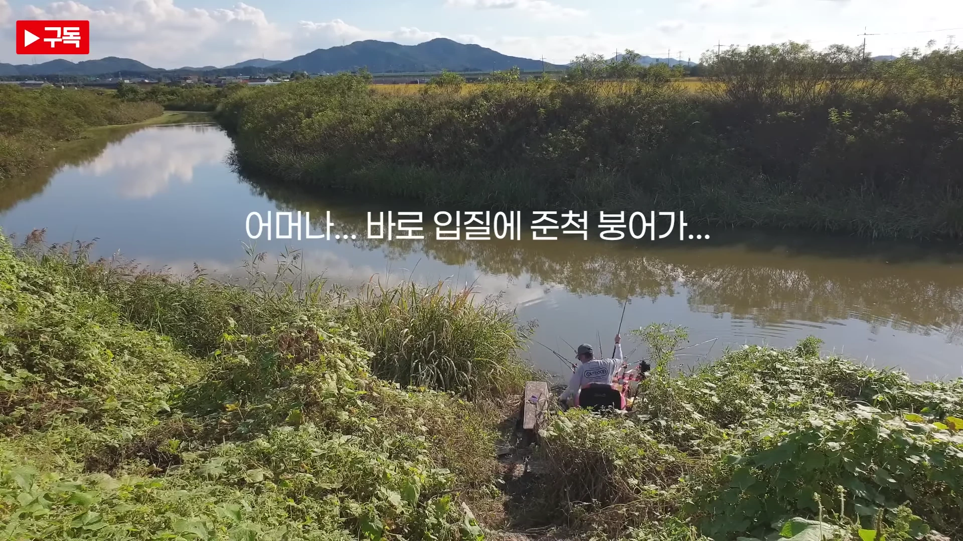 푸른 수풀로 둘러싸인 조용한 수로변에서 낚시 의자에 앉아 낚싯대를 드리운 낚시인의 뒷모습이 보이고 화면 중앙에는 '어머나... 바로 입질에 준척 붕어가...'라는 자막이 적혀 있습니다.