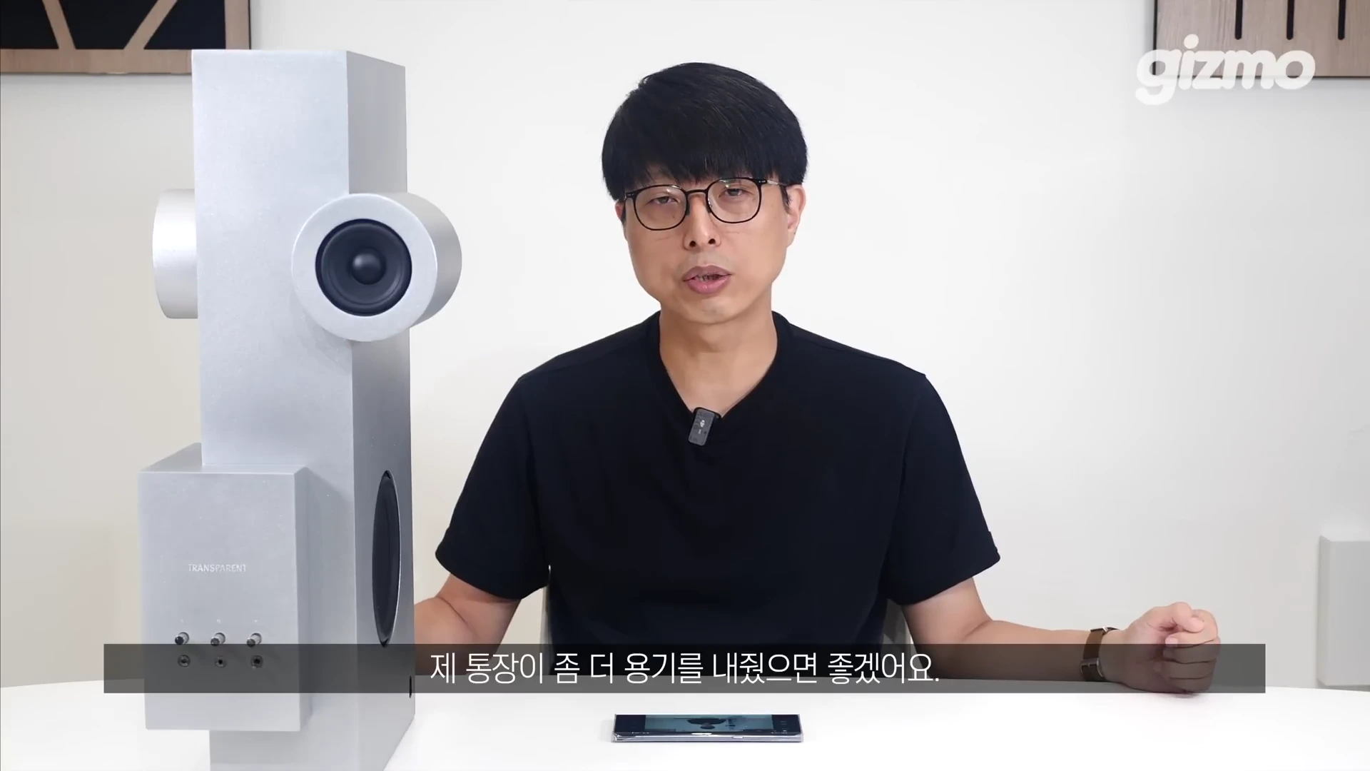 안경을 쓴 남성이 흰색 테이블 위에 놓인 금속 재질의 수직형 스피커 옆에서 이야기를 나누고 있다.