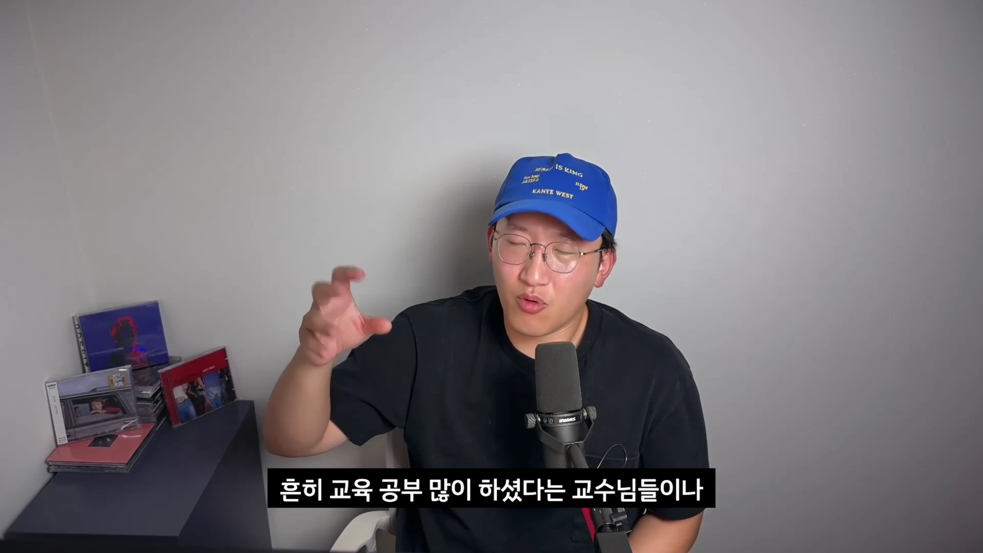 파란색 모자를 쓰고 안경을 쓴 남성이 마이크 앞에 앉아 손동작을 하며 이야기를 하고 있는 모습