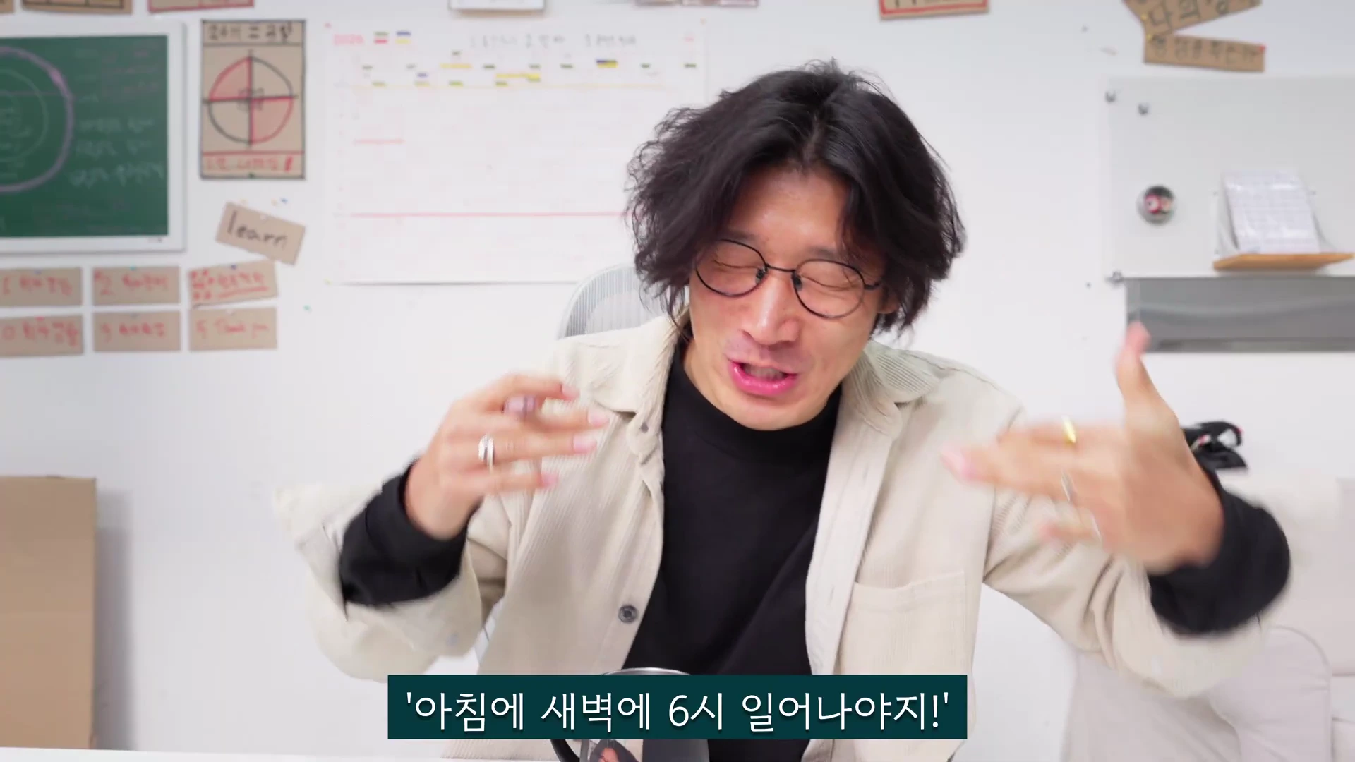 안경을 쓴 남성이 밝은 실내에서 손짓하며 무언가를 열정적으로 설명하고 있다.