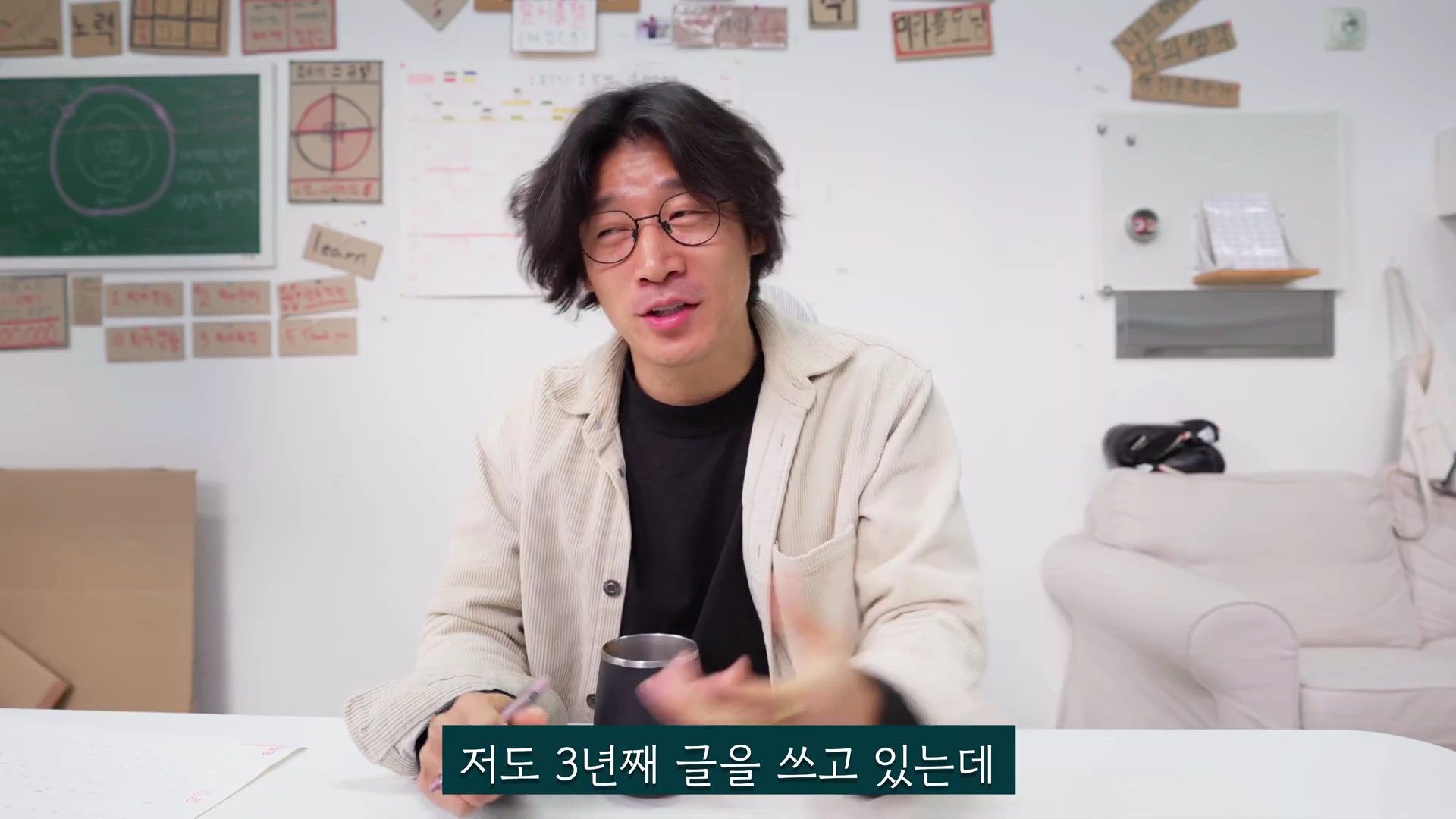 안경을 쓴 남성이 밝은 실내에서 이야기를 나누고 있으며, 배경 벽에는 여러 메모지와 게시판이 붙어 있다.