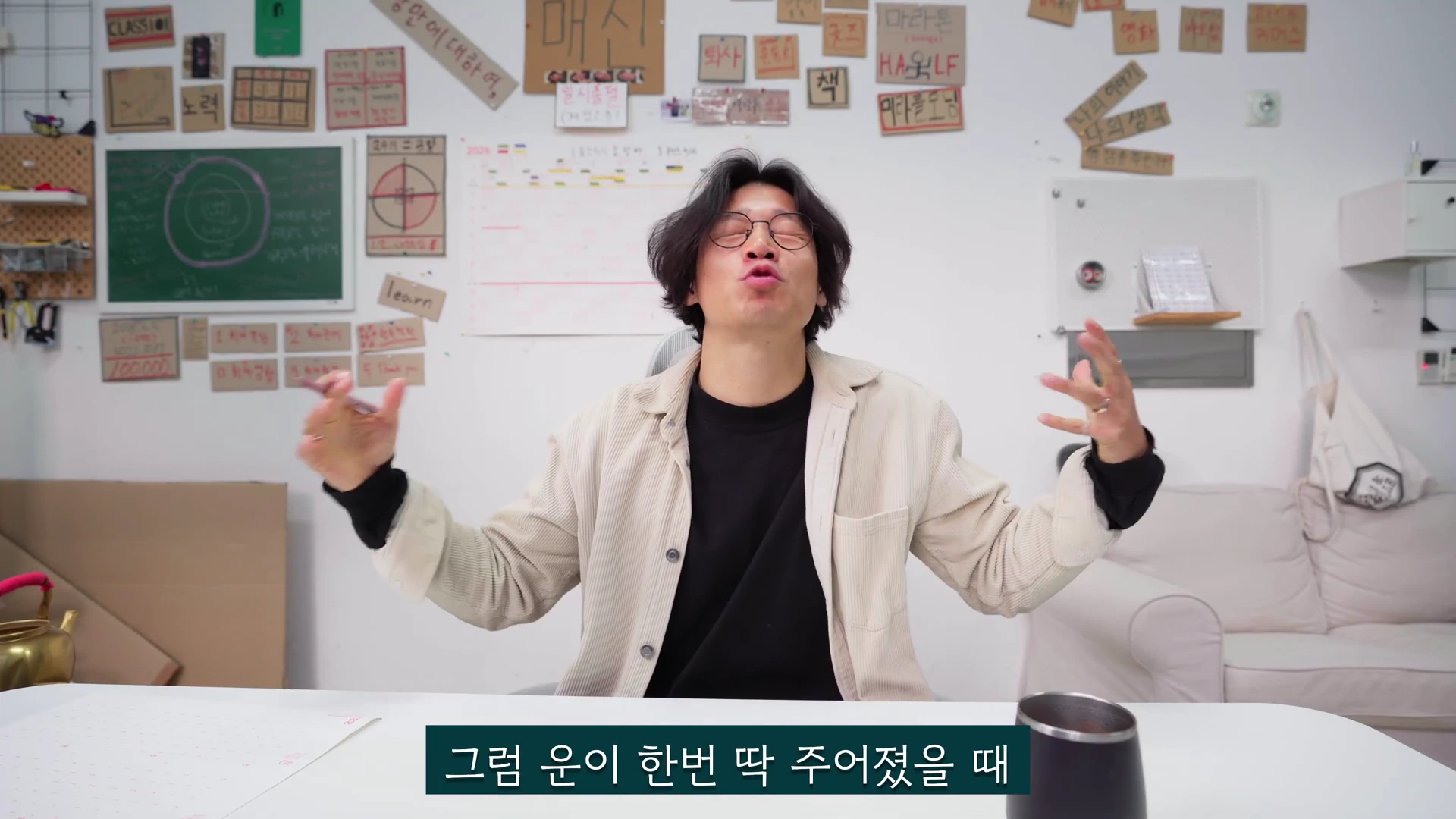 안경을 쓴 남성이 흰색 셔츠를 입고 의자에 앉아 양손을 펼치며 이야기하는 모습이 담긴 영상 캡처 화면이며, 배경의 흰 벽에는 다양한 주제가 적힌 메모지가 부착되어 있다.