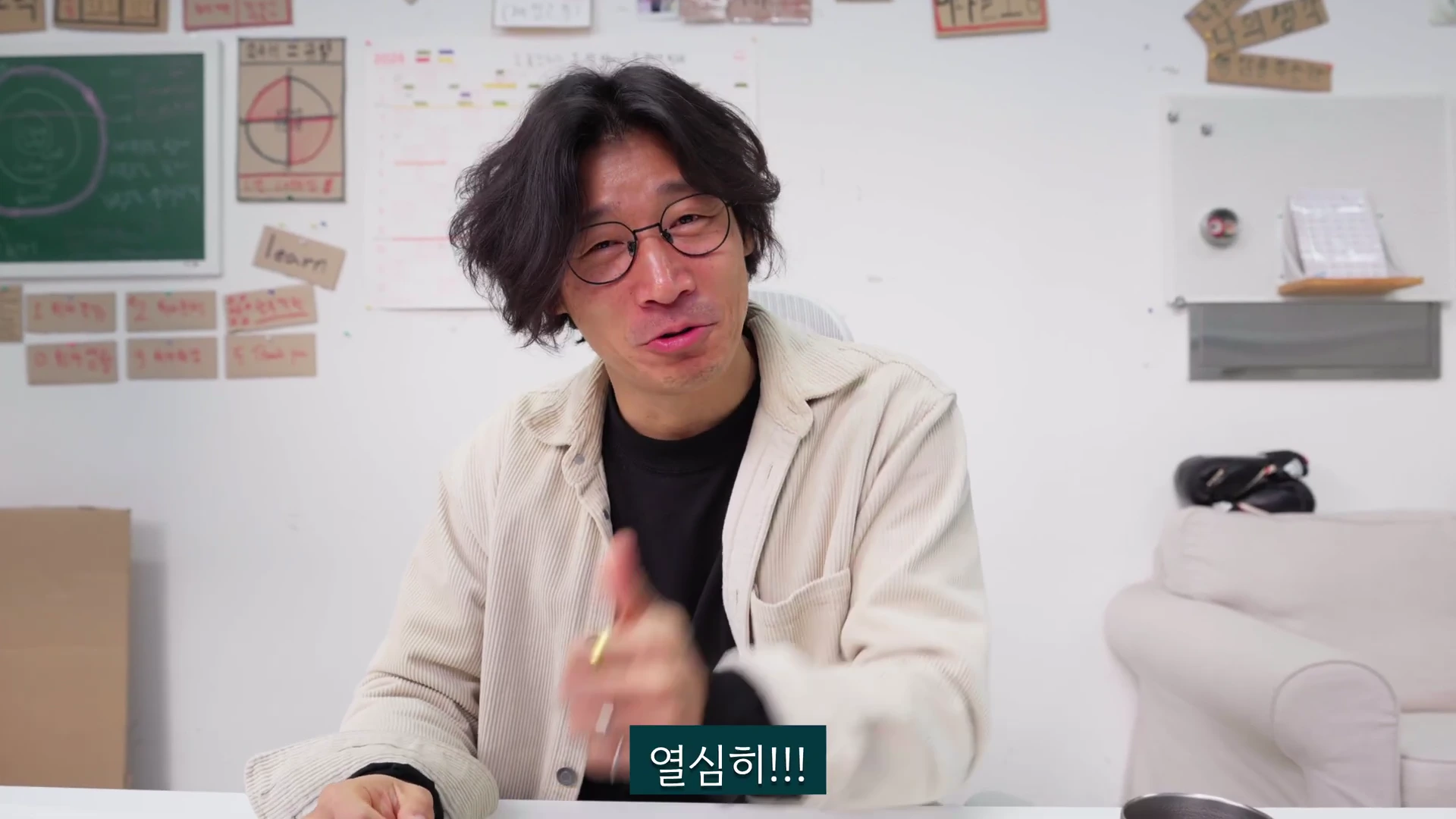 안경을 쓴 중년 남성이 흰색 벽을 배경으로 앉아 손가락으로 가리키며 열정적으로 이야기하고 있다.