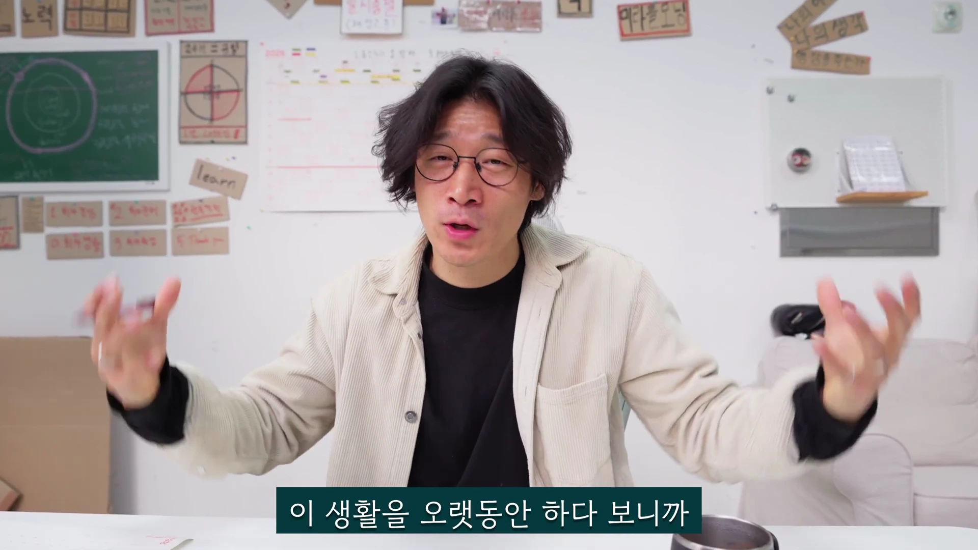 안경을 쓴 남성이 흰 벽을 배경으로 양손을 들어 제스처를 하며 이야기를 나누고 있다.