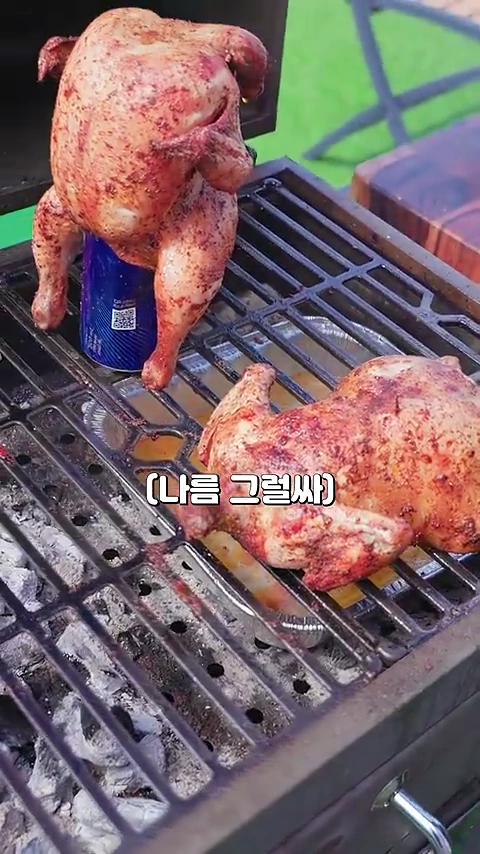 바비큐 그릴 위에 비어캔을 끼워 세운 통닭 한 마리와 그 옆에 일반 직화 방식으로 굽고 있는 또 다른 통닭이 놓여 있는 모습입니다.