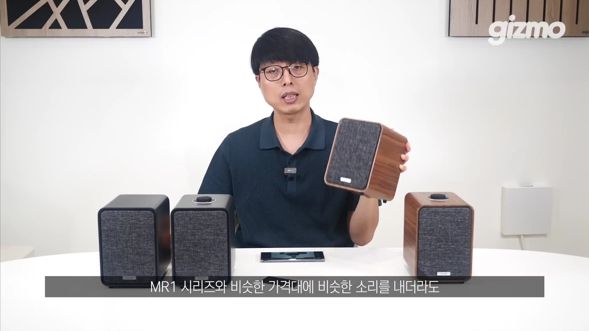 안경을 쓴 남성이 밝은 배경의 스튜디오 테이블에서 한 손으로 나무 질감의 스피커를 들어 보이고 있으며, 앞쪽 테이블에는 검은색 스피커 두 대와 스마트폰이 놓여 있습니다.