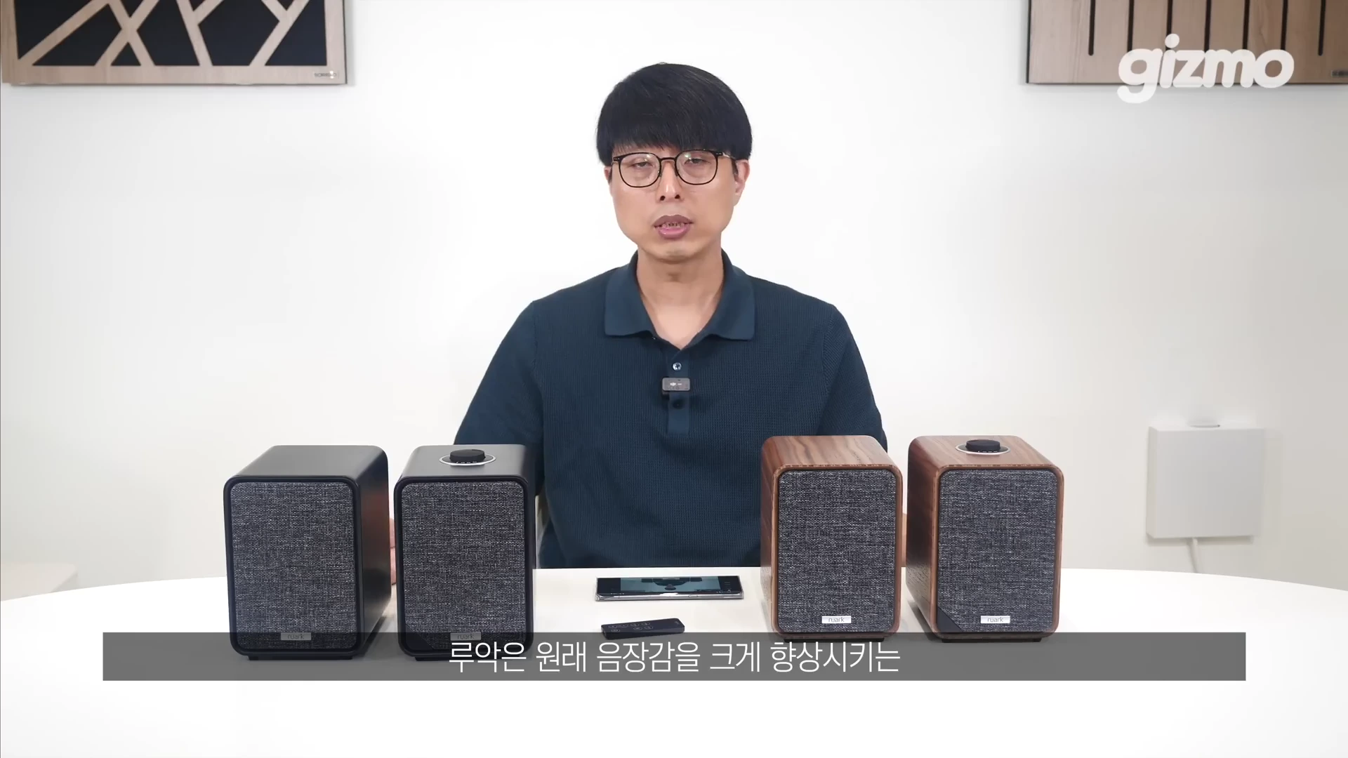 안경을 쓴 남성이 밝은 스튜디오에서 책상 위에 나란히 놓인 두 가지 색상의 스피커 쌍을 앞에 두고 이야기하고 있다.