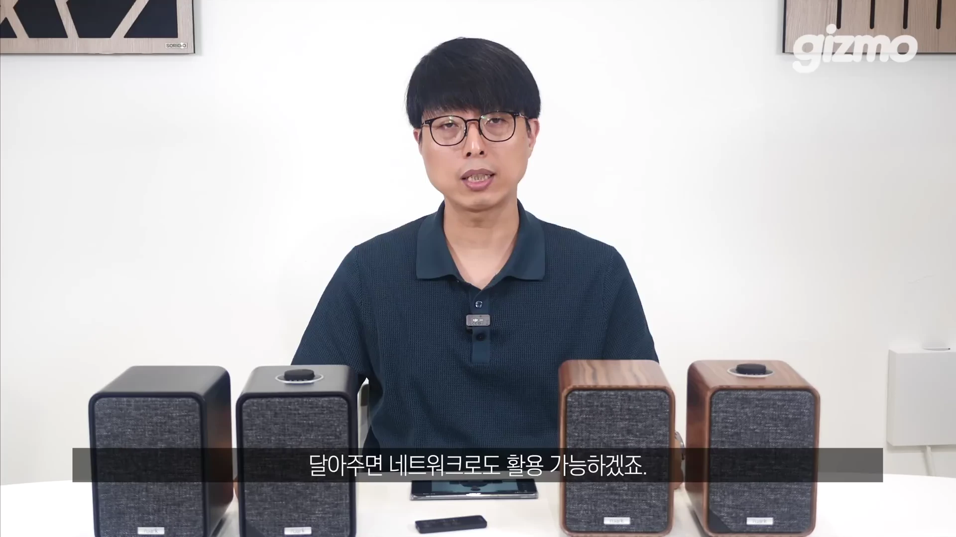 흰색 배경의 스튜디오에서 안경을 쓴 남성이 탁자 위에 놓인 검은색과 나무 무늬의 스피커들을 앞에 두고 이야기하고 있다.