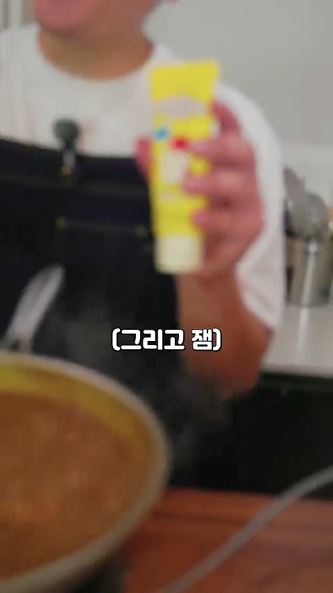 흰색 티셔츠와 검은색 앞치마를 입은 사람이 한 손에 노란색 튜브형 잼을 들고 보여주는 모습과 그 아래 끓고 있는 카레가 담긴 프라이팬이 보인다.