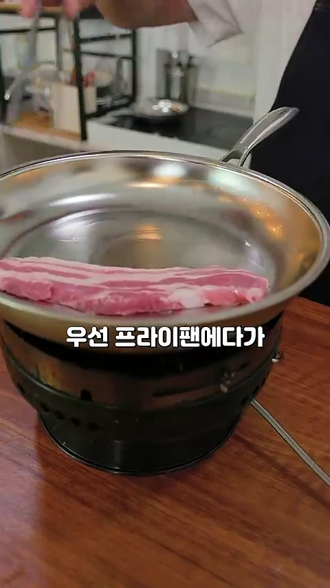 주방에서 스테인리스 프라이팬에 두툼한 생삼겹살 한 줄을 올려 굽기 시작하는 모습.