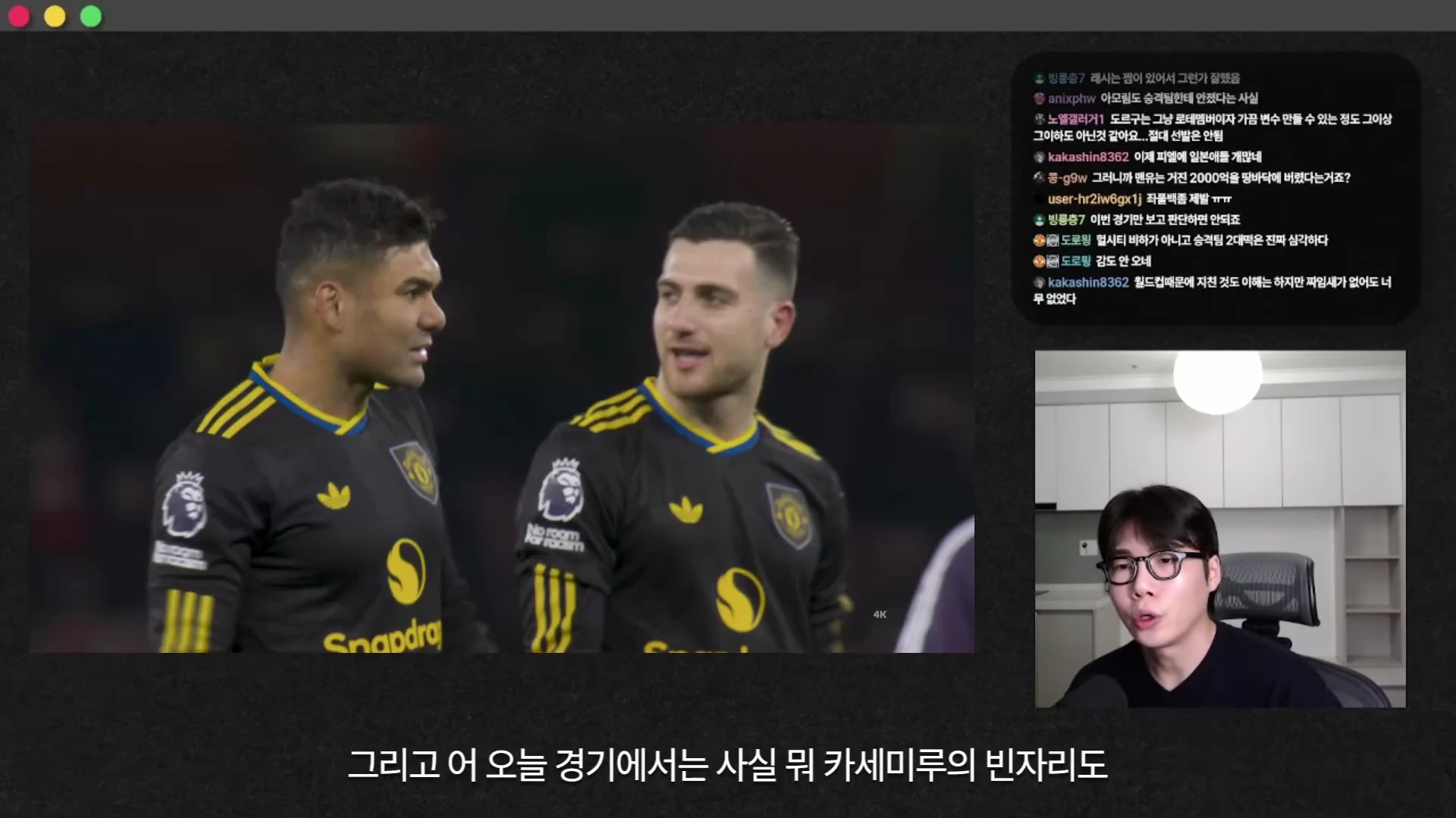 축구 경기 중 경기장에 서 있는 검은색 유니폼을 입은 두 선수와, 이를 해설하는 안경 쓴 남성이 나타난 방송 화면