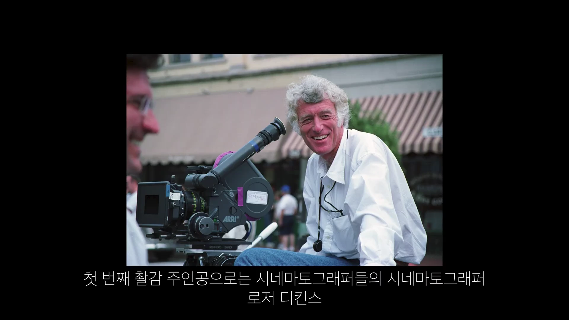 흰 셔츠를 입은 은발의 남성이 영화 촬영용 카메라 옆에서 밝게 웃고 있는 모습
