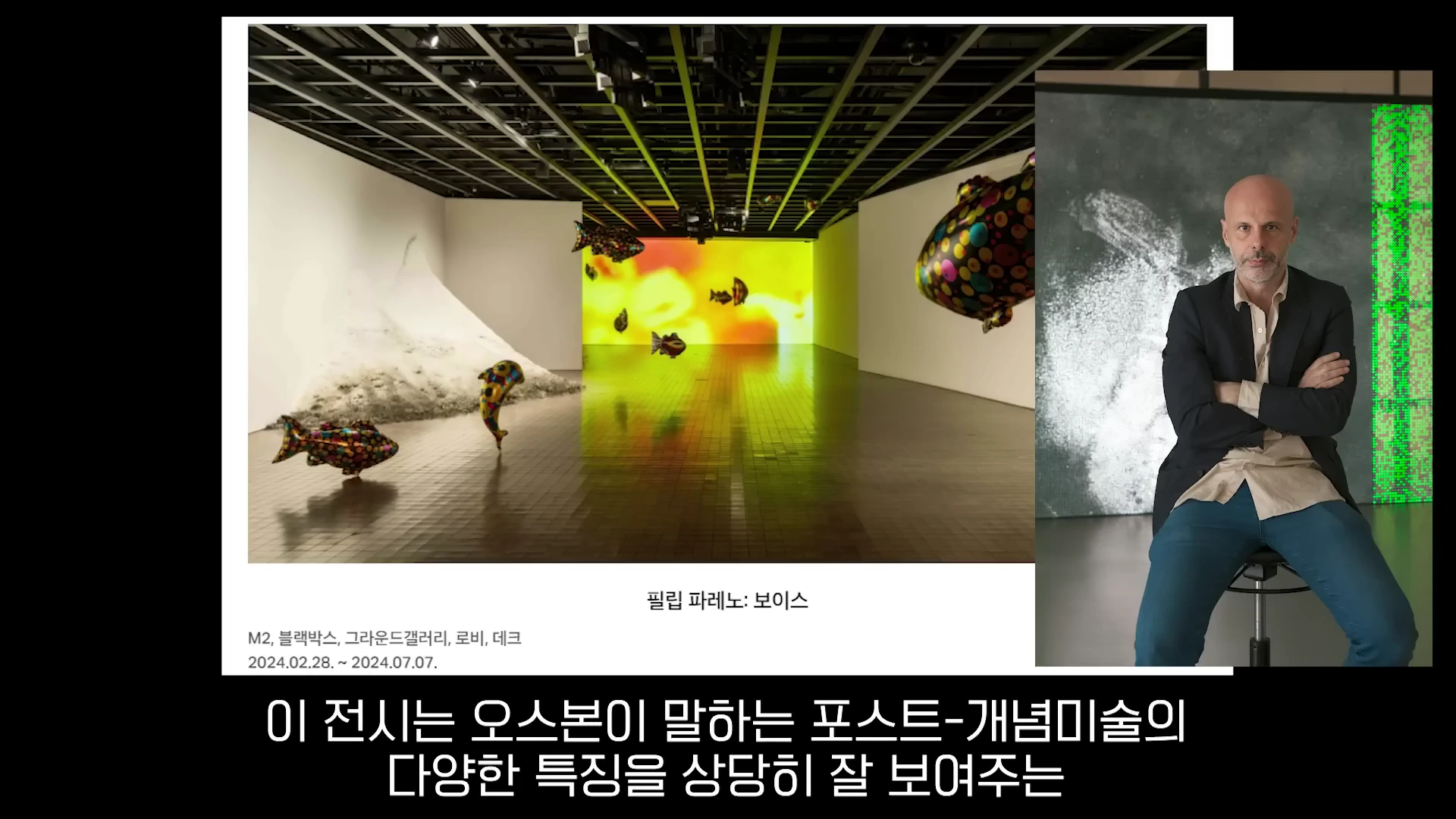 화면 왼쪽에는 물고기 모양 풍선이 떠다니는 전시 공간이 보이고, 오른쪽에는 팔짱을 끼고 의자에 앉아 있는 대머리의 남성이 화면 앞에 위치해 있다.