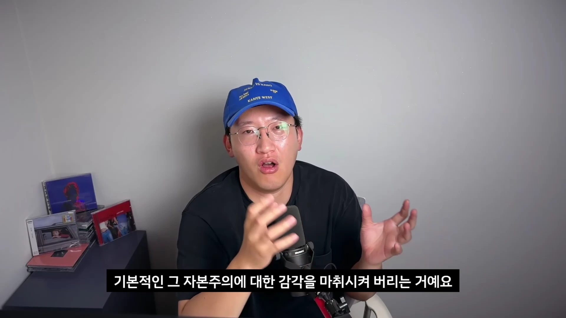 파란색 야구 모자와 안경을 쓴 남성이 마이크 앞에서 손짓하며 이야기를 하고 있다. 배경에는 앨범 자켓들이 놓여 있다.