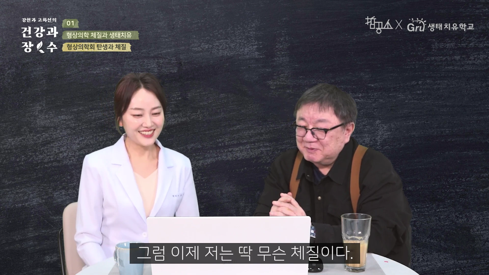 흰색 가운을 입은 여성과 검은색 셔츠를 입고 안경을 쓴 남성이 테이블 앞에 앉아 대화하는 모습의 스튜디오 영상 화면