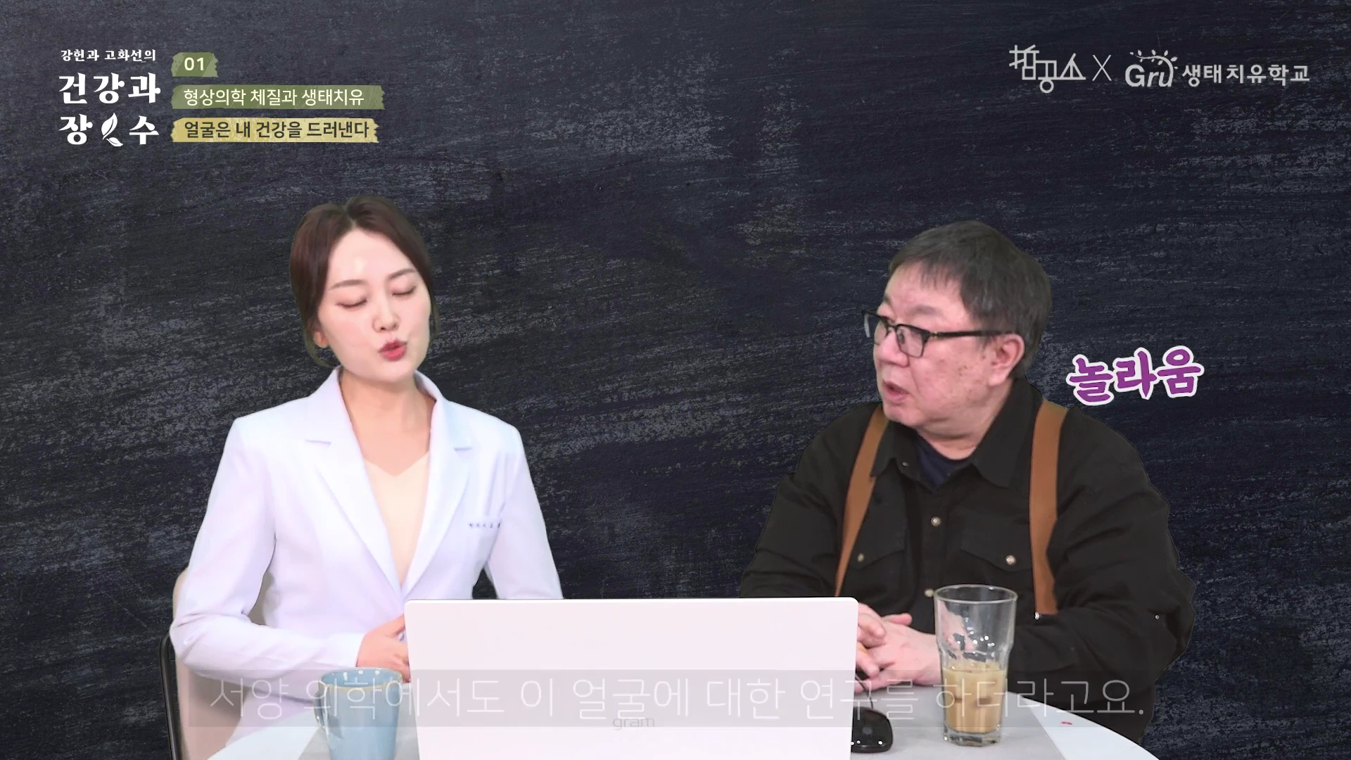 스튜디오 배경에서 하얀색 재킷을 입은 여성과 검은색 셔츠를 입은 남성이 테이블에 앉아 대화를 나누는 모습.
