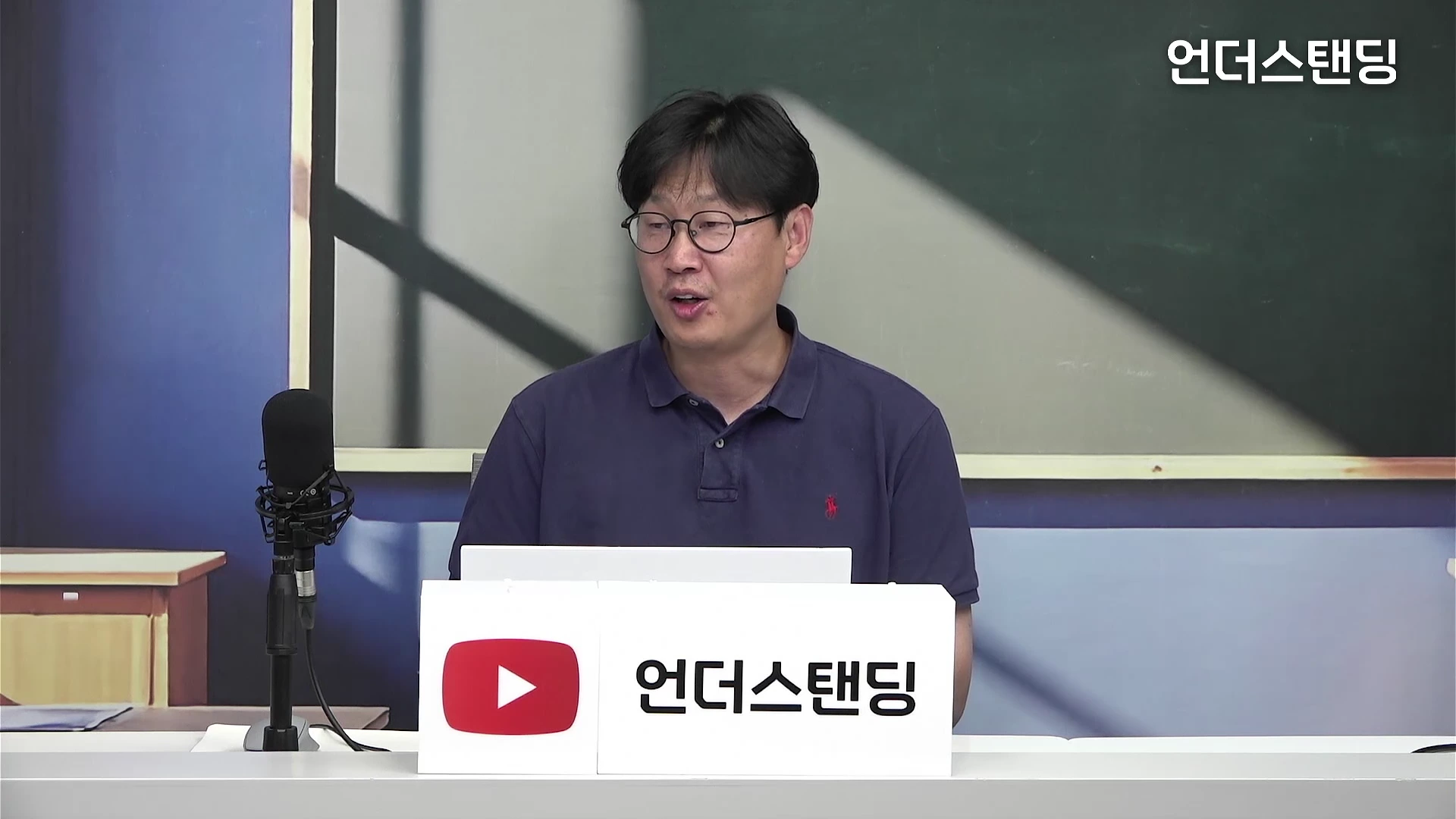안경을 쓴 중년 남성이 스튜디오 책상 앞에 앉아 마이크를 앞에 두고 이야기를 나누고 있다.