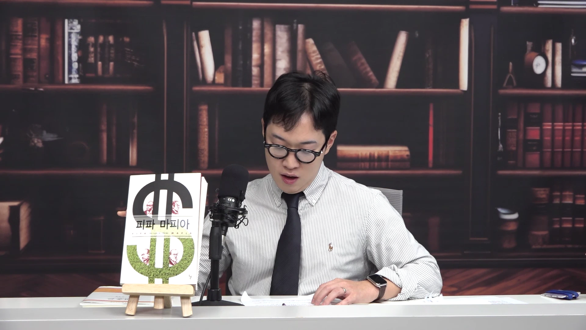 서재 배경 앞에서 안경을 쓴 남성이 '피파 마피아'라는 제목의 책을 손으로 가리키며 이야기하고 있는 스튜디오 장면