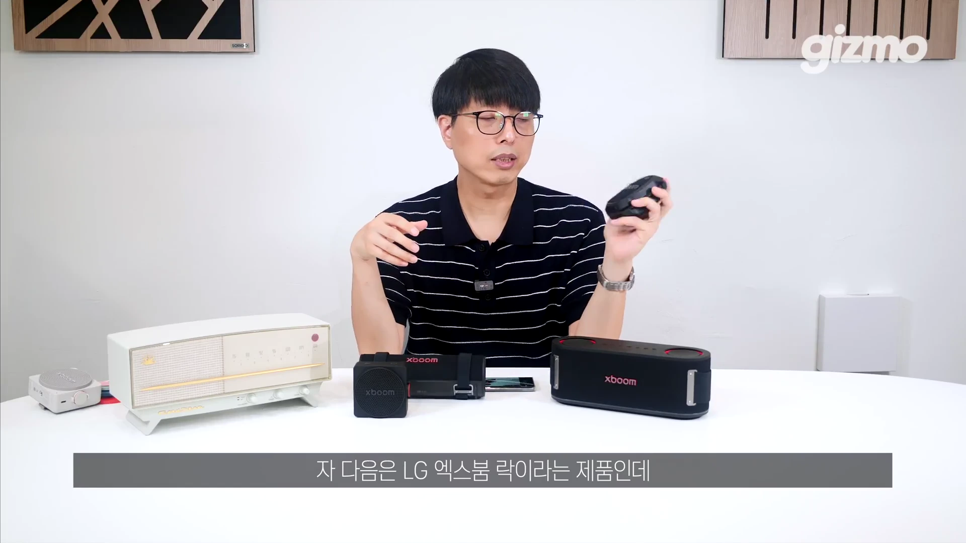안경을 쓴 남성이 테이블 위에 놓인 LG 엑스붐 스피커 모델들을 소개하고 있으며, 손에는 작은 휴대용 스피커를 들고 설명하고 있다.