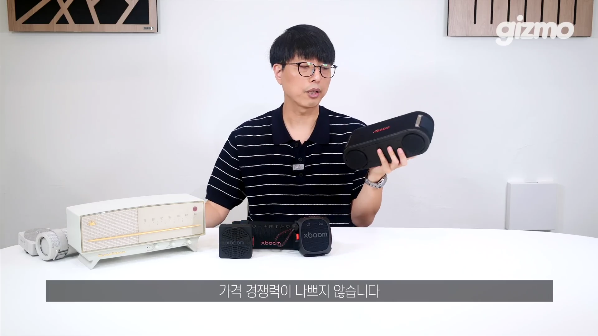 안경을 쓴 남성이 흰색 테이블 위에 놓인 다양한 LG 엑스붐 스피커 제품들을 보여주며 설명하고 있다.