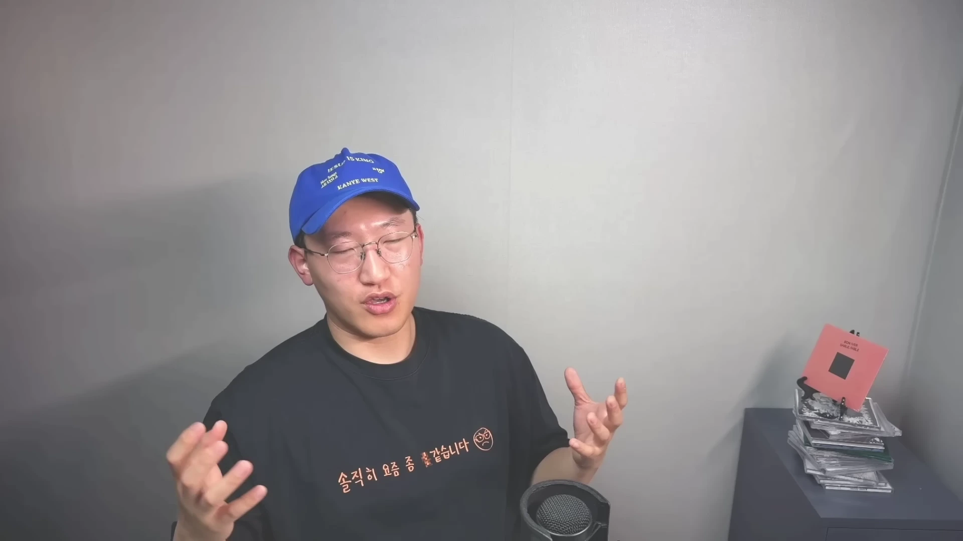 파란색 야구 모자를 쓰고 검은색 티셔츠를 입은 안경 쓴 남성이 마이크 앞에서 손동작을 하며 이야기하고 있다.