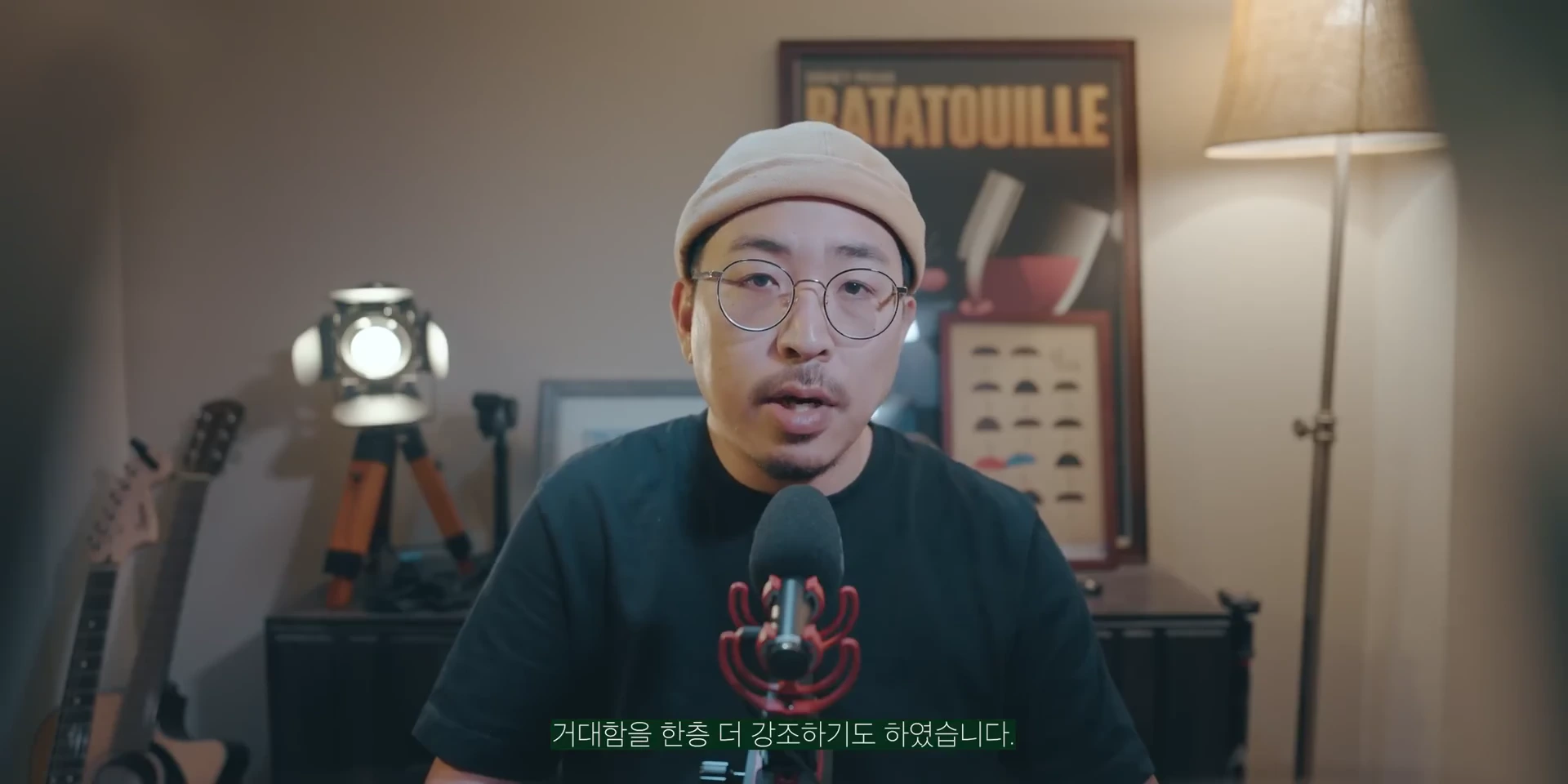 비니를 쓰고 안경을 쓴 남성이 마이크 앞에 앉아 대화하고 있는 모습이 담긴 정면 스튜디오 촬영 화면