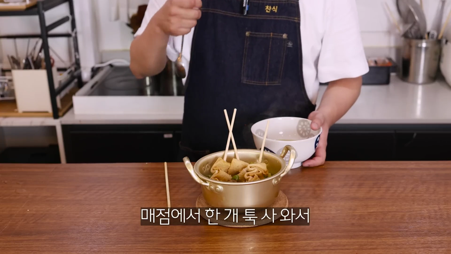앞치마를 입은 요리사가 어묵 꼬치가 담긴 양은 냄비를 앞에 두고 숟가락으로 국물을 뜨고 있는 모습
