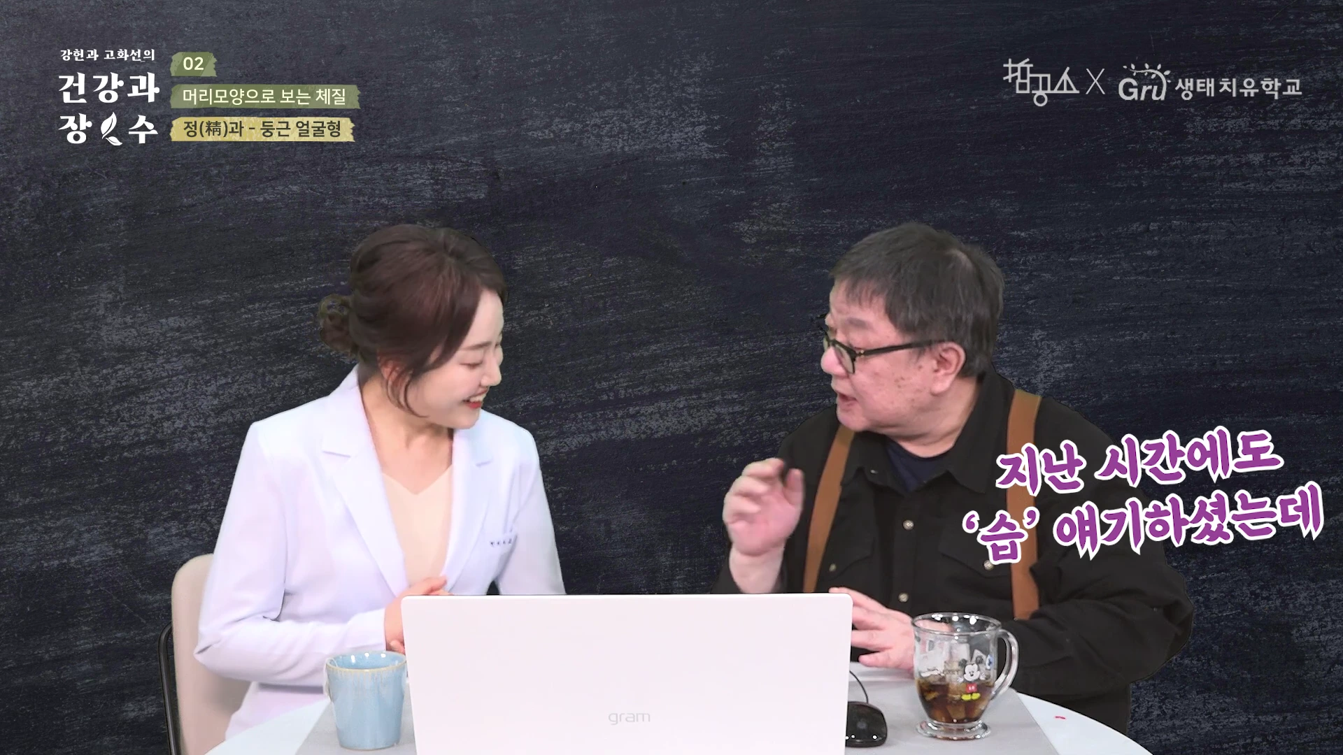 흰 가운을 입은 여성과 검은 셔츠를 입은 남성이 책상에 앉아 대화를 나누고 있는 스튜디오 인터뷰 장면