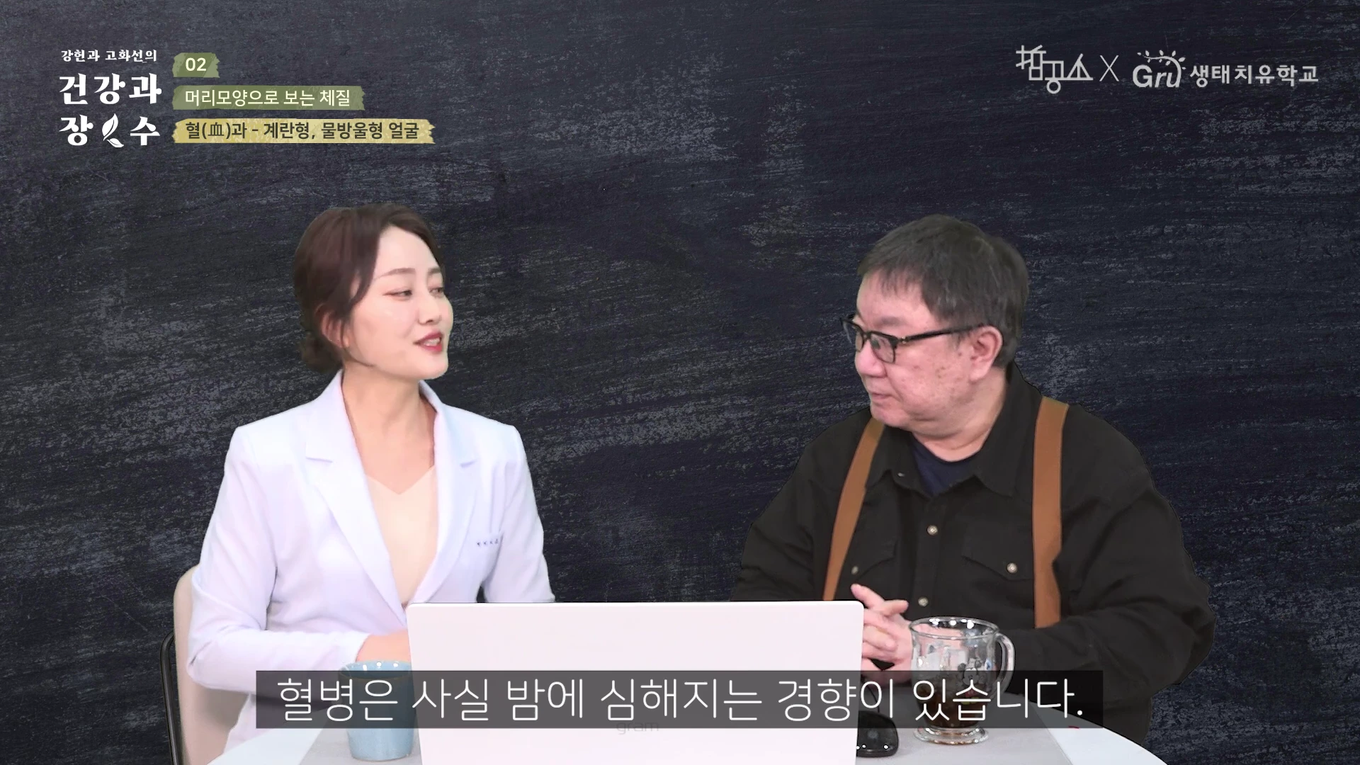 흰 가운을 입은 여성과 안경을 쓴 남성이 검은 배경의 스튜디오에서 대화를 나누고 있으며, 화면 하단에는 자막이 떠 있습니다.