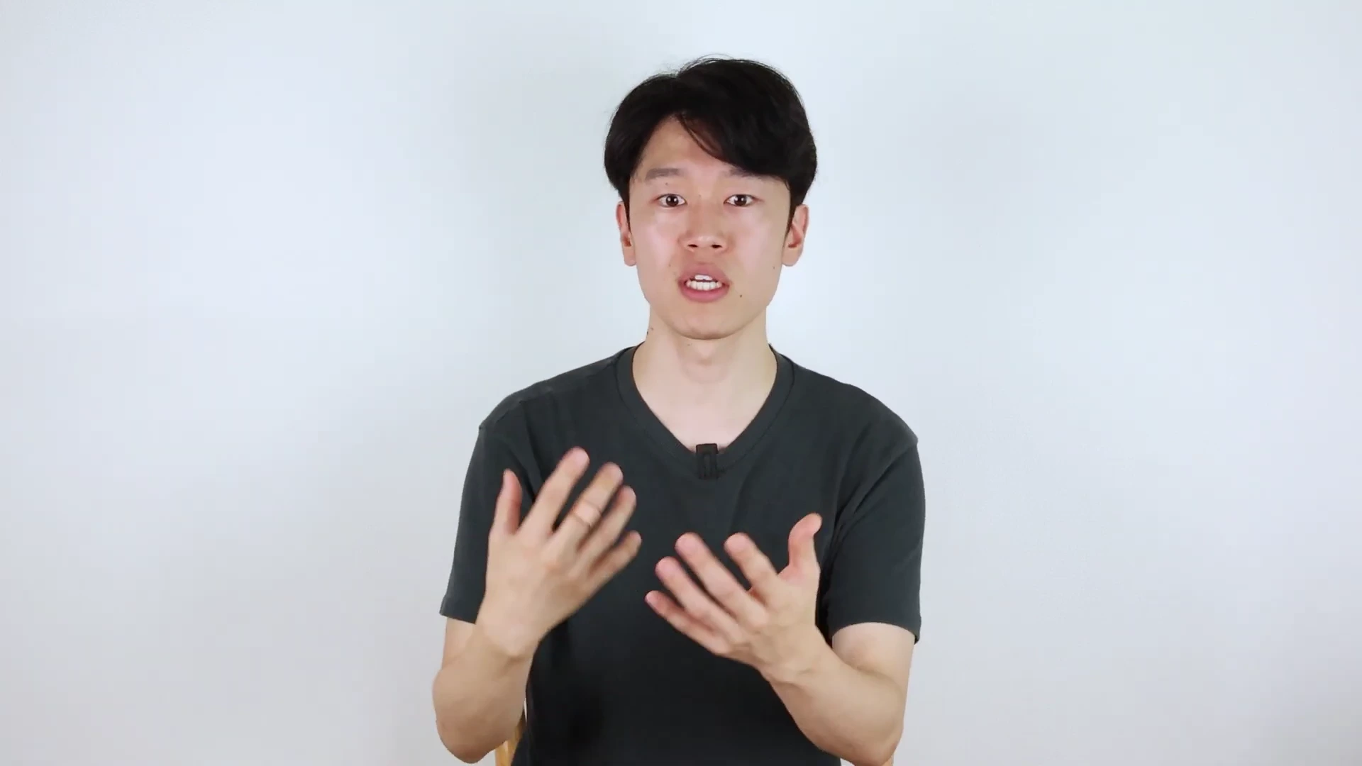 짙은 녹색 브이넥 티셔츠를 입은 젊은 남성이 흰색 배경 앞에서 손동작을 하며 무언가를 설명하고 있다.