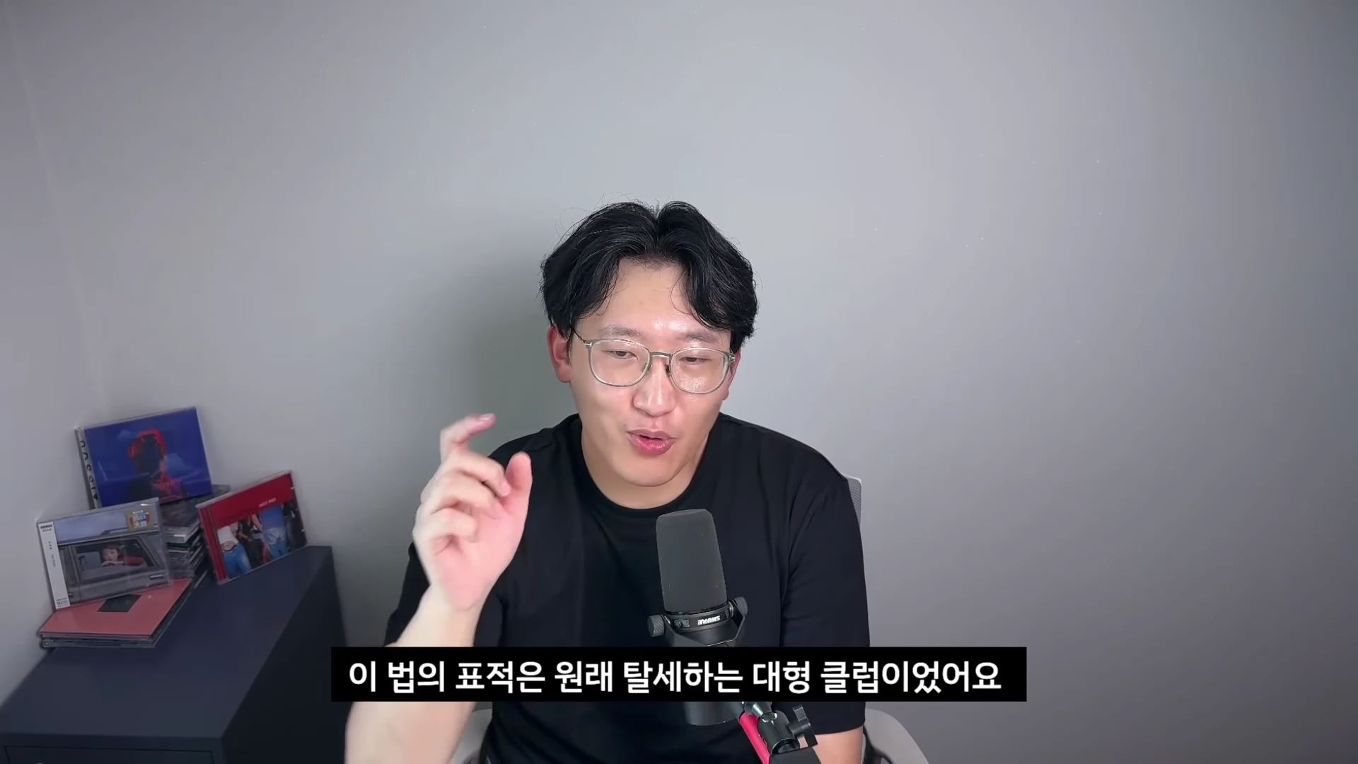 A man with glasses and a black t-shirt is sitting in front of a microphone, speaking to the camera in a studio setting, with a video caption overlay in Korean.