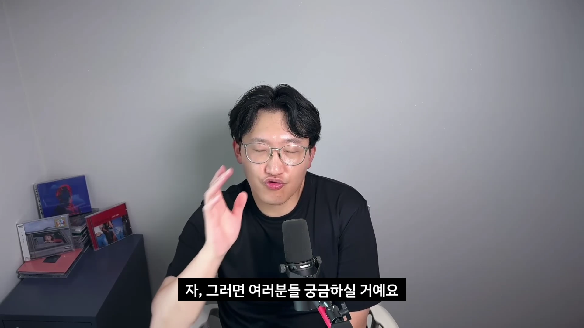 검은색 반소매 티셔츠를 입고 안경을 쓴 남성이 마이크 앞에 앉아 손을 들어 설명하는 모습입니다.