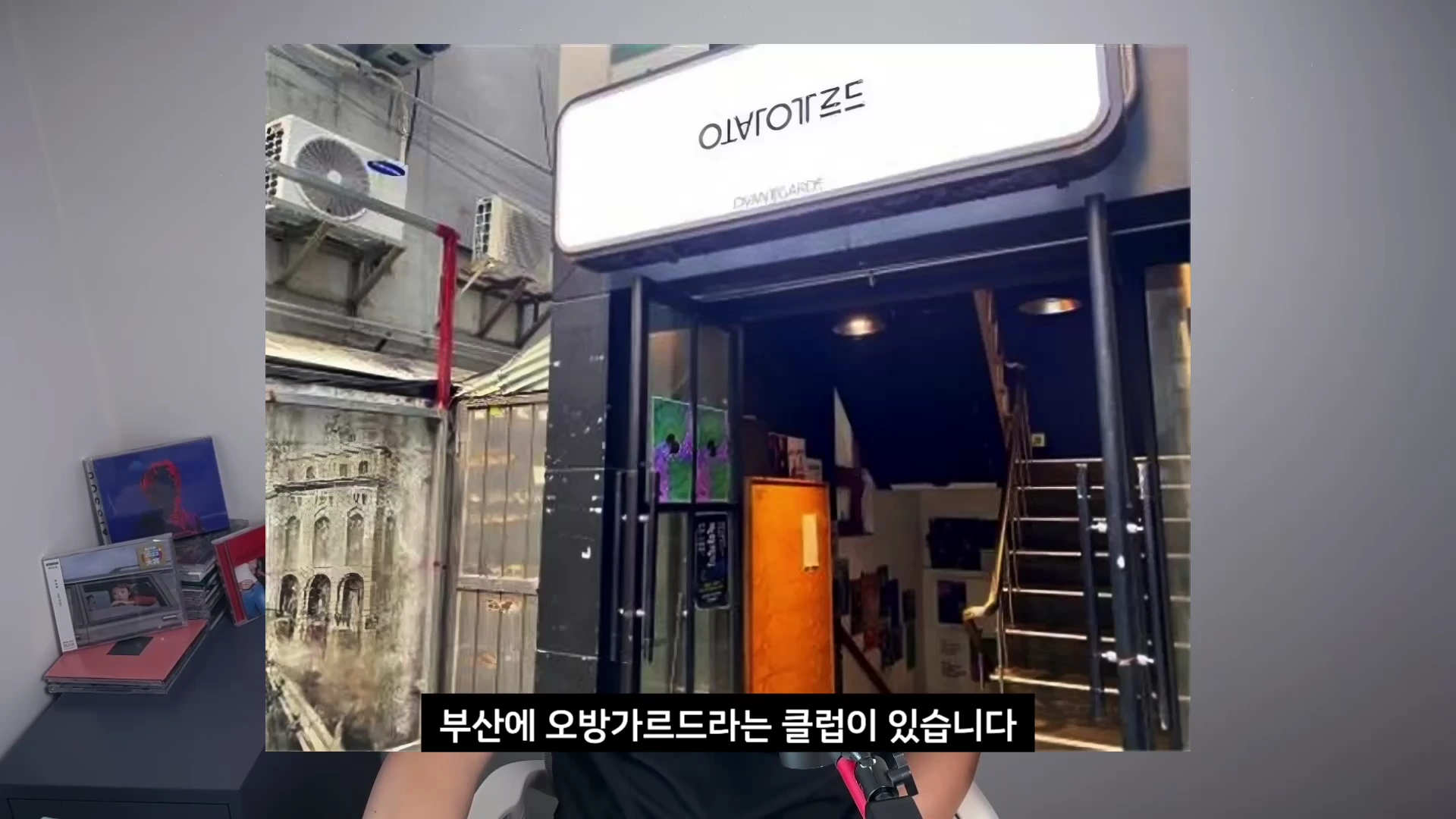 흰색 간판이 걸린 지하 라이브 클럽 입구와 아래로 이어지는 계단이 보이며, 화면 하단에는 한국어 자막이 삽입되어 있습니다.