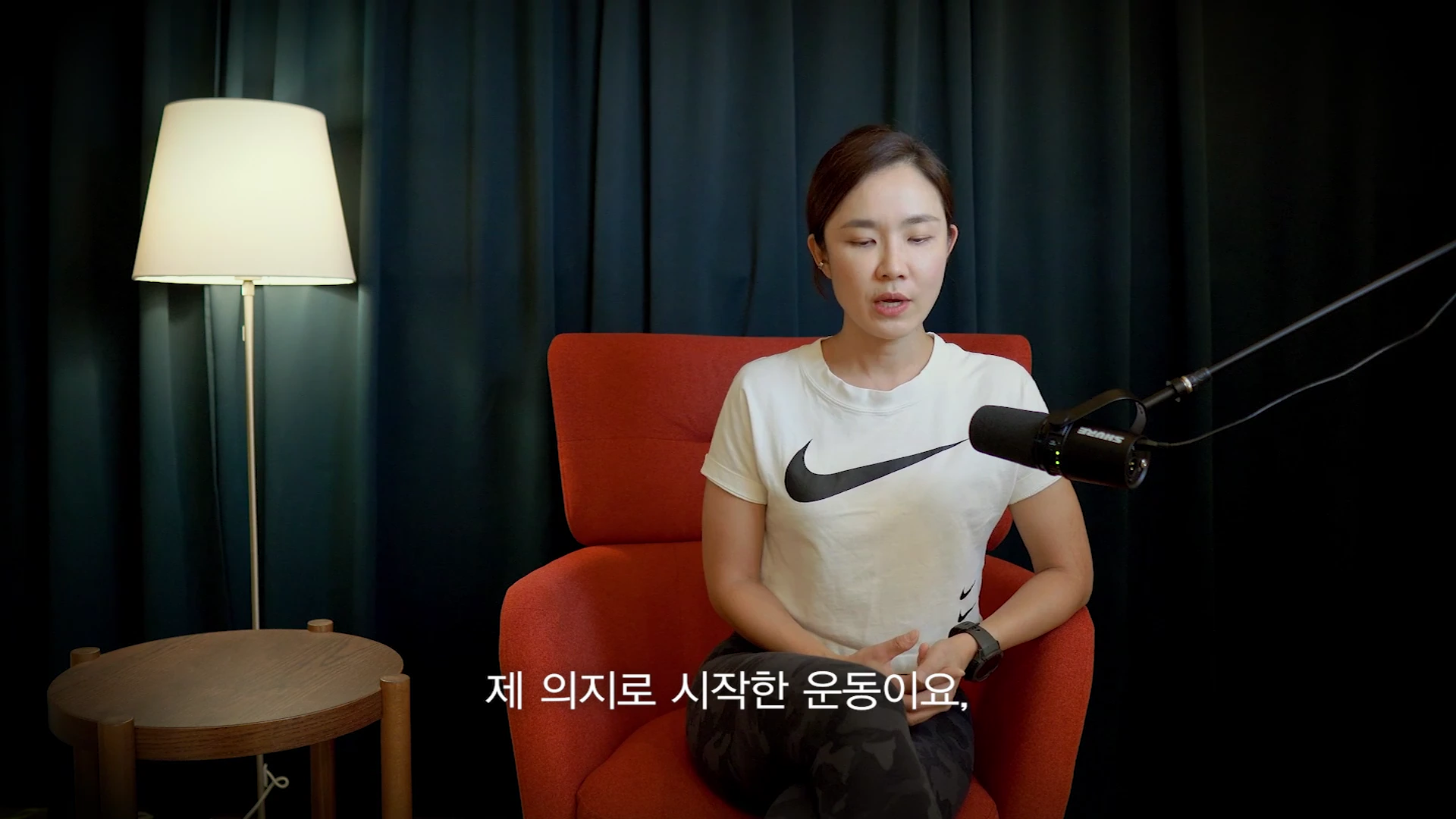 흰색 나이키 티셔츠를 입은 여성이 붉은색 의자에 앉아 검은색 스탠드 마이크 앞에서 인터뷰를 진행하고 있으며, 화면 하단에는 '제 의지로 시작한 운동이요'라는 자막이 표시되어 있습니다.