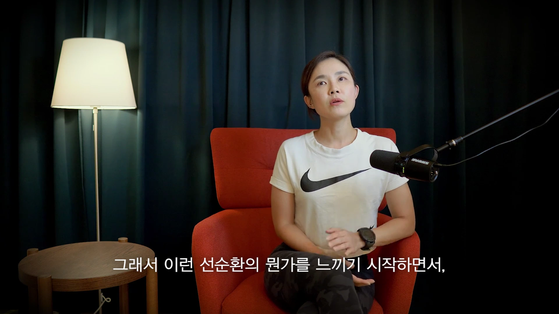 흰색 나이키 티셔츠를 입은 여성이 빨간 의자에 앉아 검은색 마이크 앞에서 인터뷰를 하고 있으며, 왼쪽에는 노란색 전등이 켜져 있습니다.
