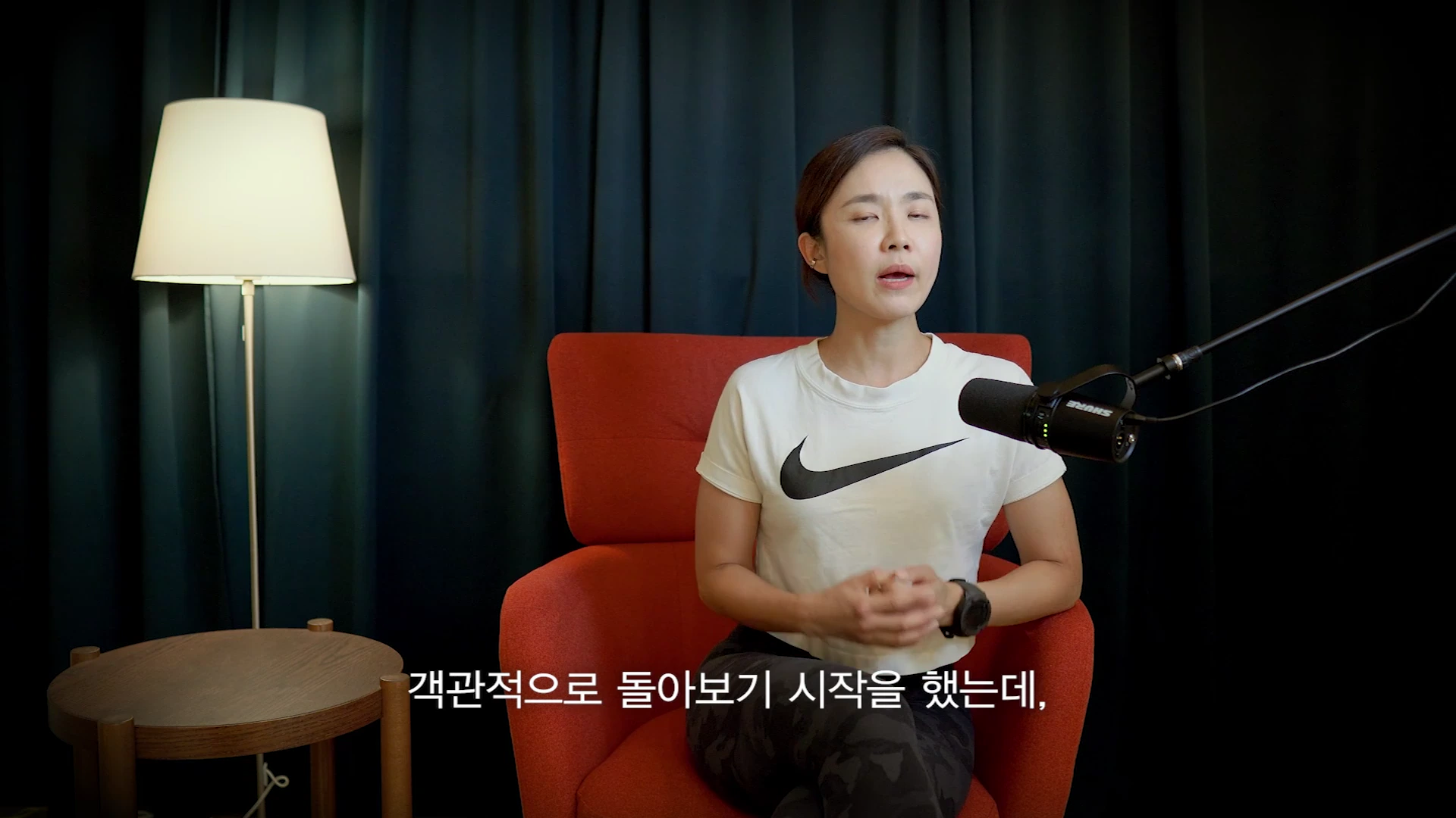 흰색 반팔 티셔츠를 입은 한 여성이 어두운 배경의 스튜디오에서 붉은색 의자에 앉아 인터뷰를 하고 있으며 앞에 마이크가 놓여 있습니다.
