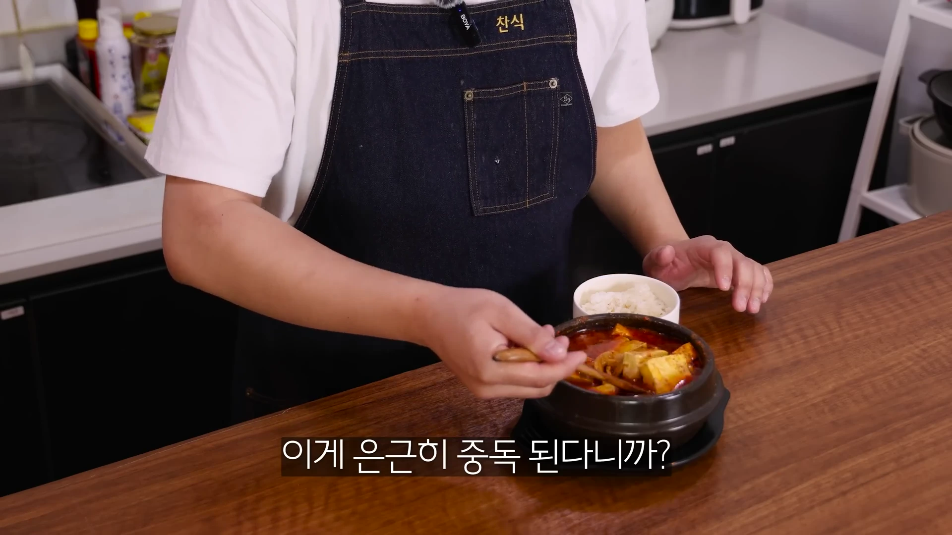 앞치마를 두른 요리사가 뚝배기에 담긴 찌개를 숟가락으로 젓고 있는 모습이며 화면 하단에 자막이 떠 있습니다.
