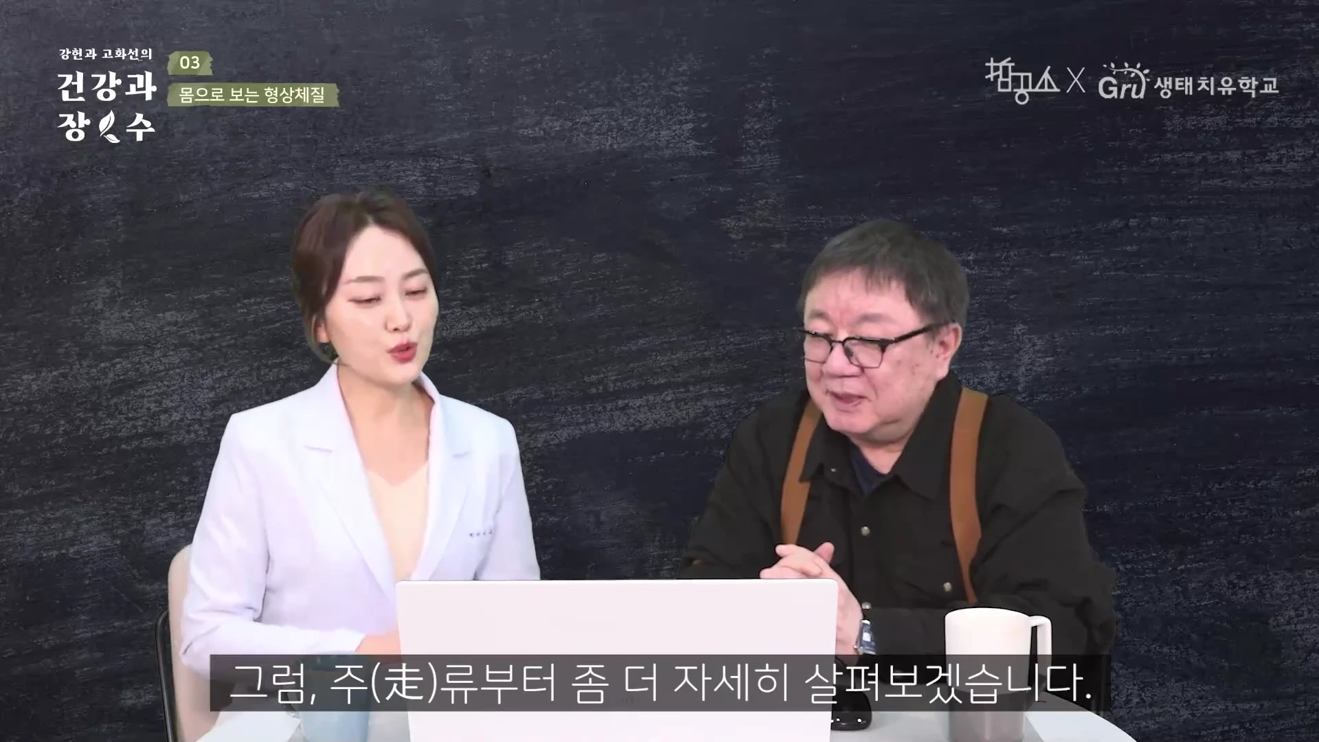 두 명의 진행자가 탁자에 앉아 대화를 나누고 있는 스튜디오 배경의 영상 캡처 화면이며, 하단에는 '그럼, 주류부터 좀 더 자세히 살펴보겠습니다'라는 자막이 표시되어 있습니다.