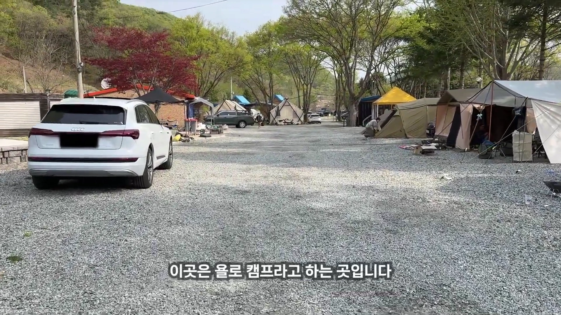 흰색 SUV가 주차된 넓은 자갈밭 캠핑장에 여러 형태의 텐트들이 설치되어 있고 나무들이 주변을 둘러싸고 있는 야외 풍경