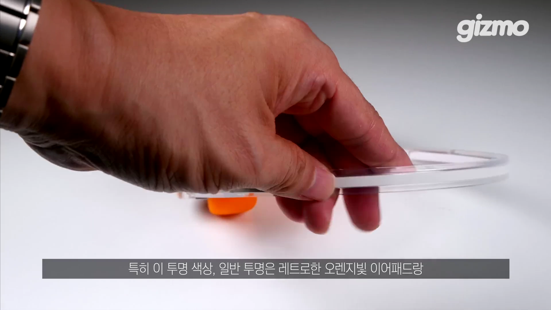 한 손으로 투명한 헤드폰 프레임을 잡고 있는 모습이며, 아래에 주황색 이어패드가 일부 보입니다.