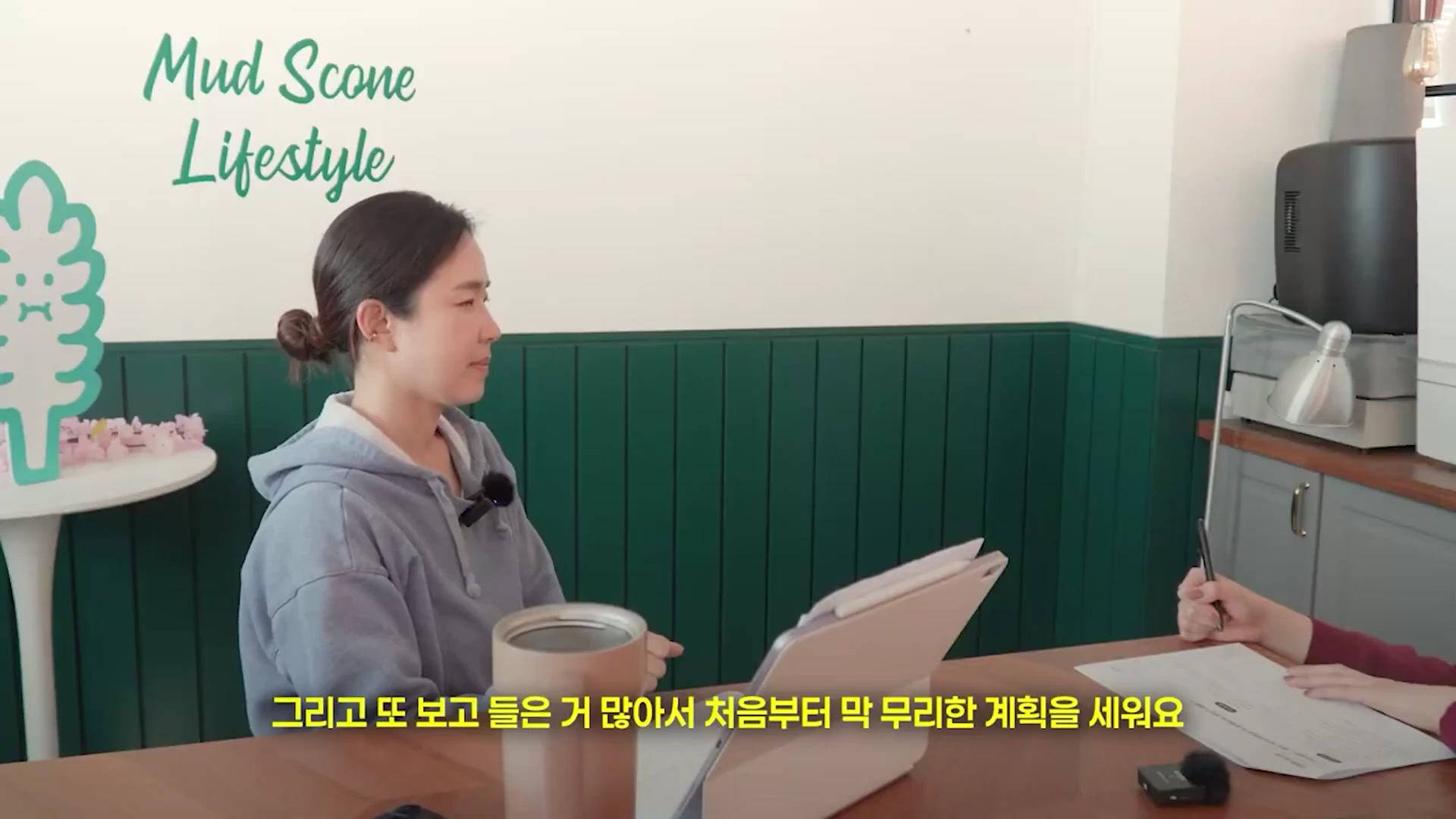 실내에서 후드티를 입은 여성이 테이블에 앉아 인터뷰를 진행하고 있으며, 화면 하단에는 '그리고 또 보고 들은 거 많아서 처음부터 막 무리한 계획을 세워요'라는 한국어 자막이 표시되어 있습니다.