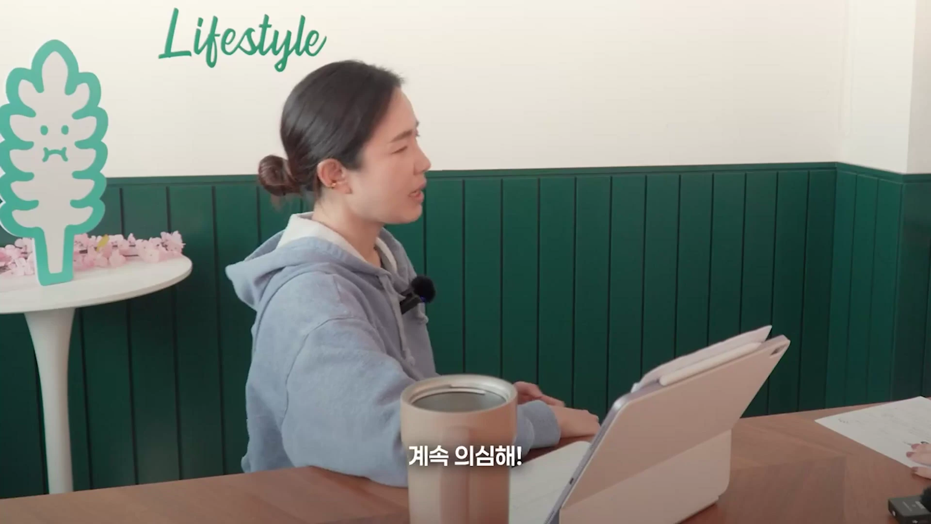 실내에서 후드티를 입은 여성이 책상 앞에 앉아 이야기하는 모습