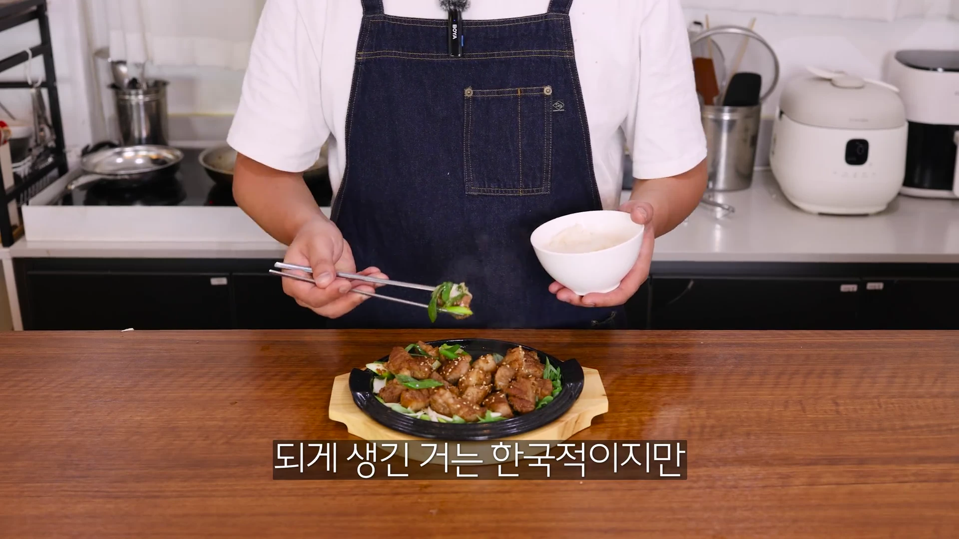 앞치마를 두른 요리사가 젓가락으로 구운 대파를 집어 올리고 한 손에는 밥공기를 든 채, 철판 위에 놓인 먹음직스러운 목살 구이를 보여주고 있는 주방의 모습.