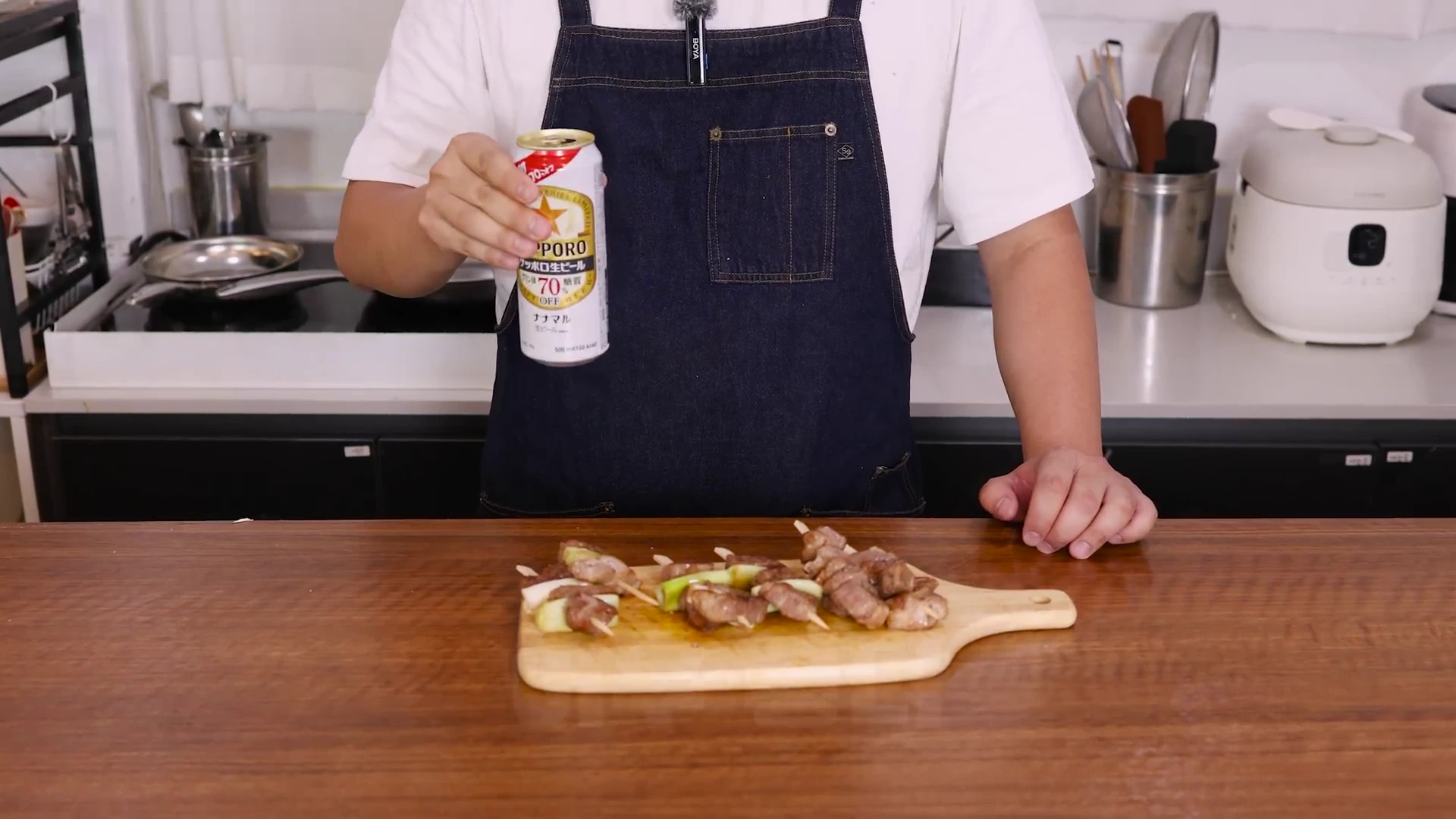 앞치마를 두른 요리사가 한 손에는 캔맥주를 들고 다른 한 손으로는 나무 도마 위에 올려진 돼지고기 대파 꼬치 요리를 가리키고 있다.