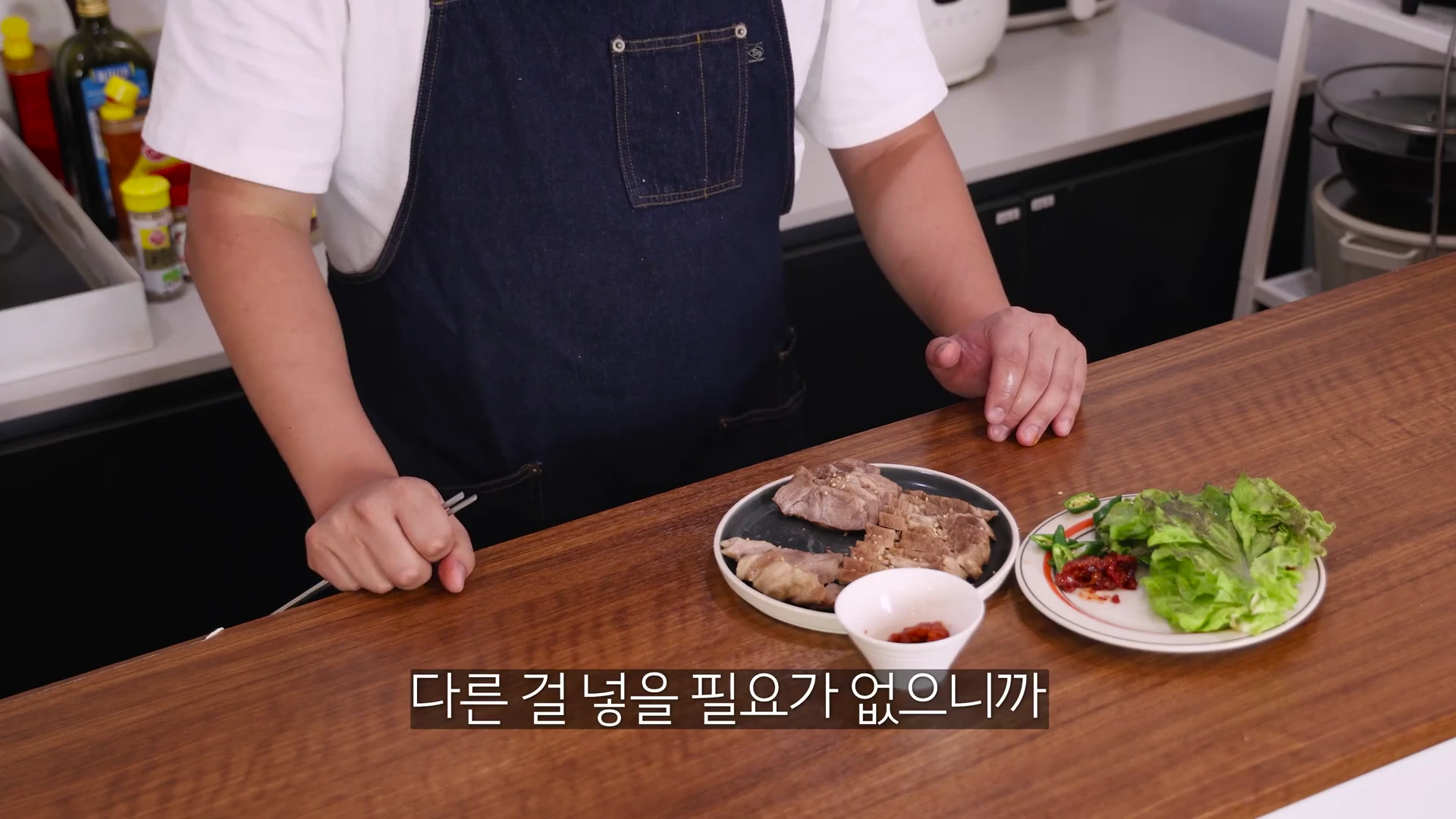 앞치마를 두른 요리사가 나무 식탁 위에 완성된 통목살 수육 접시와 쌈 채소를 놓아두고 있는 모습