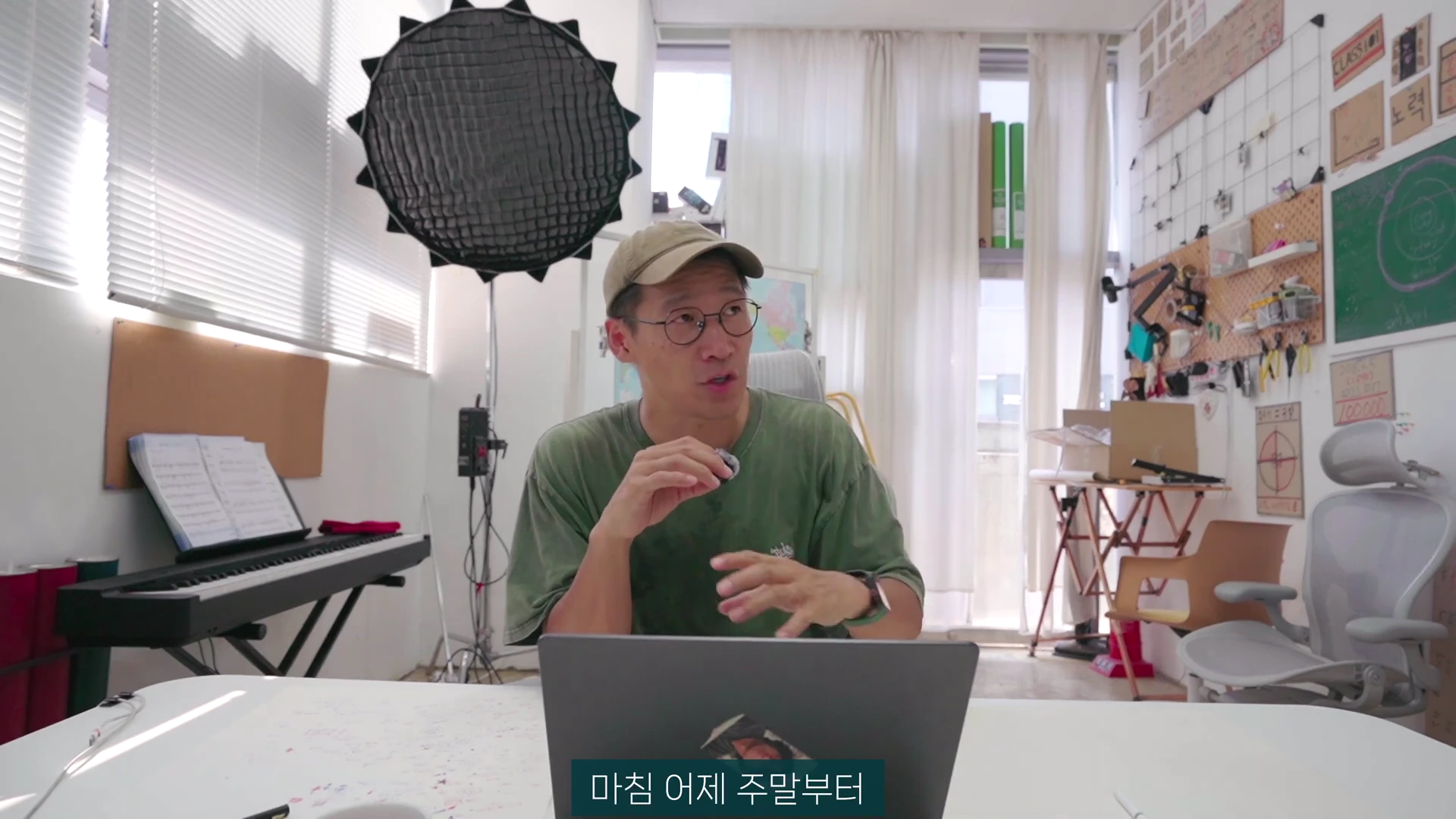 모자를 쓴 남성이 밝은 작업실에서 책상 앞에 앉아 노트북을 켜두고 이야기를 나누고 있는 모습이다.